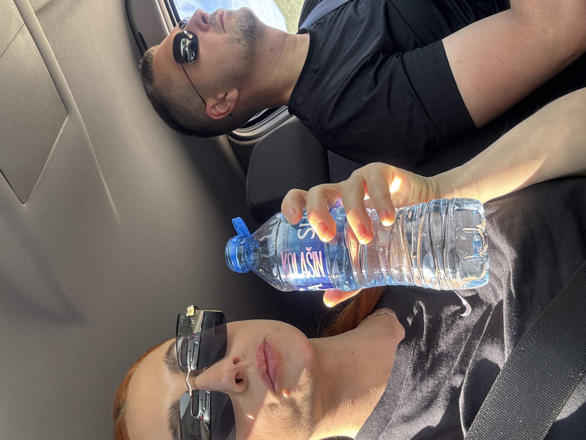 The image depicts two individuals inside a vehicle, seemingly during a car ride. The person on the right of the frame is a male wearing sunglasses and a black t-shirt. The person on the left is a female with red hair, wearing sunglasses and a black t-shirt and seatbelt. She is holding a bottle of water labeled "KOLASIN". The scene does not appear to depict Podlastva (Poljice), Montenegro. It seems to be an interior shot inside a car.