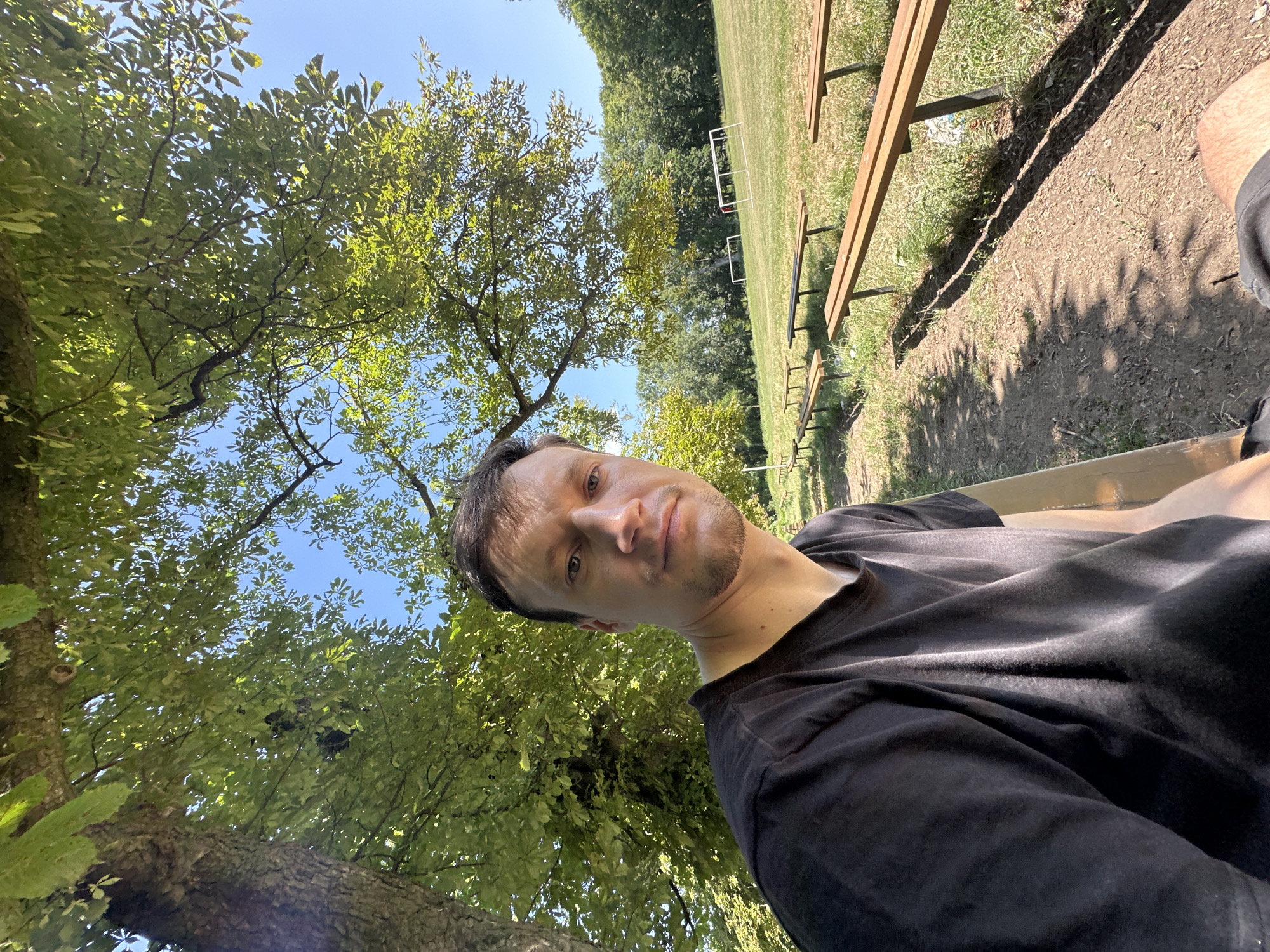 A low-angle, selfie-style shot captures a man sitting outdoors on a sunny day. He is wearing a black t-shirt and has short brown hair and a light beard. He is looking directly at the camera.
Behind him, a dirt path runs alongside several wooden benches, which appear to be seating for an outdoor sports field. The field itself is visible in the background, with white goalposts marking a soccer field. Trees with green foliage frame the scene, with sunlight filtering through the leaves. The sky is clear and blue.
The image appears to be taken in a park or recreational area. Based on the presence of a soccer field and benches, this could be a local sports ground. The location is identified as Makiv, Ukraine.