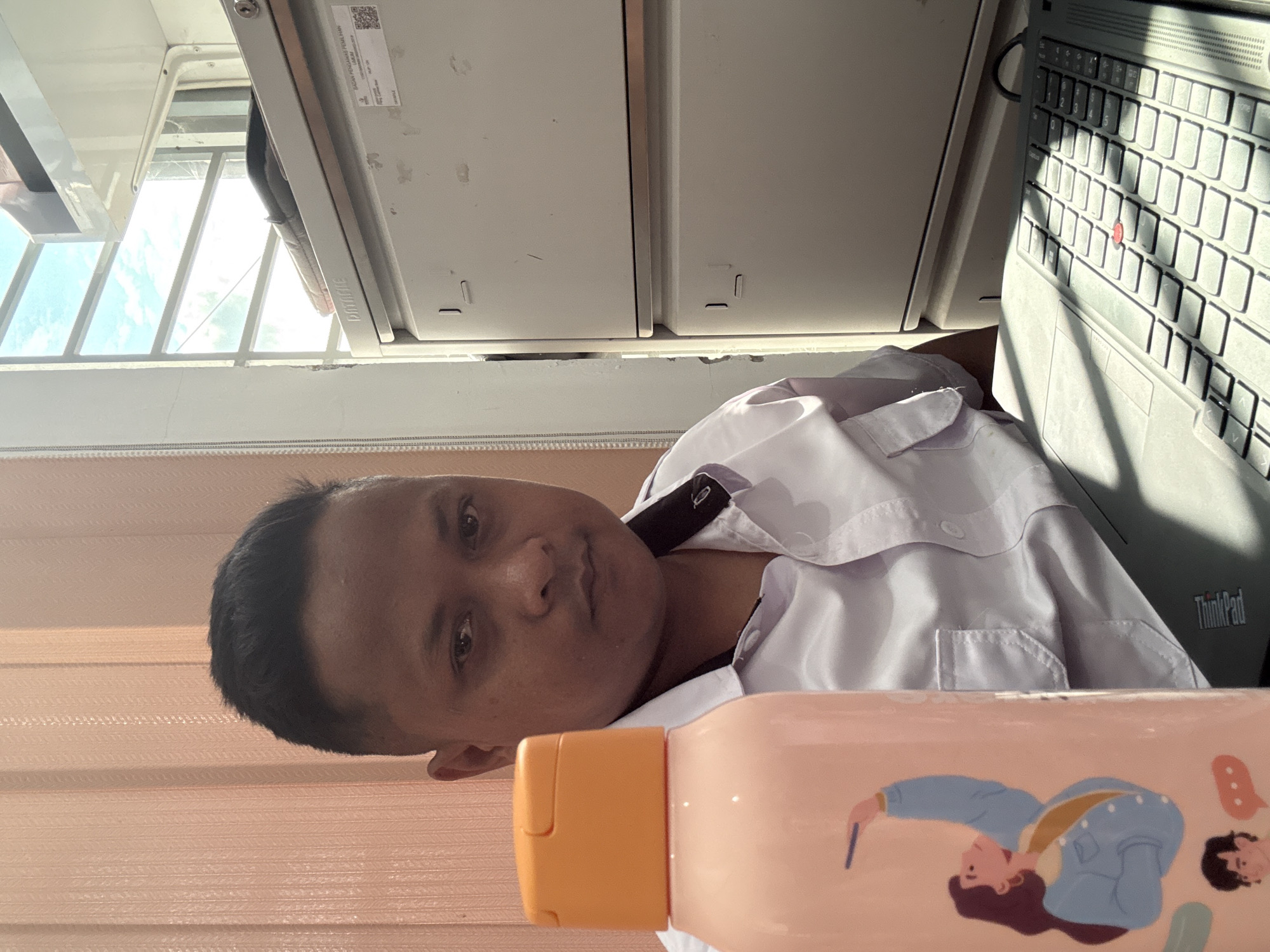 A medium shot shows a person with short dark hair and brown skin, wearing a white collared shirt, positioned indoors next to a laptop. The laptop is partially visible with a black keyboard. A peach-colored bottle with cartoon illustrations is in the foreground. In the background, there's a window with blinds, and what appears to be storage units. The person is looking directly at the camera. Based on the background and other details, it is not possible to confirm the location to be Lhokseumawe, Indonesia.