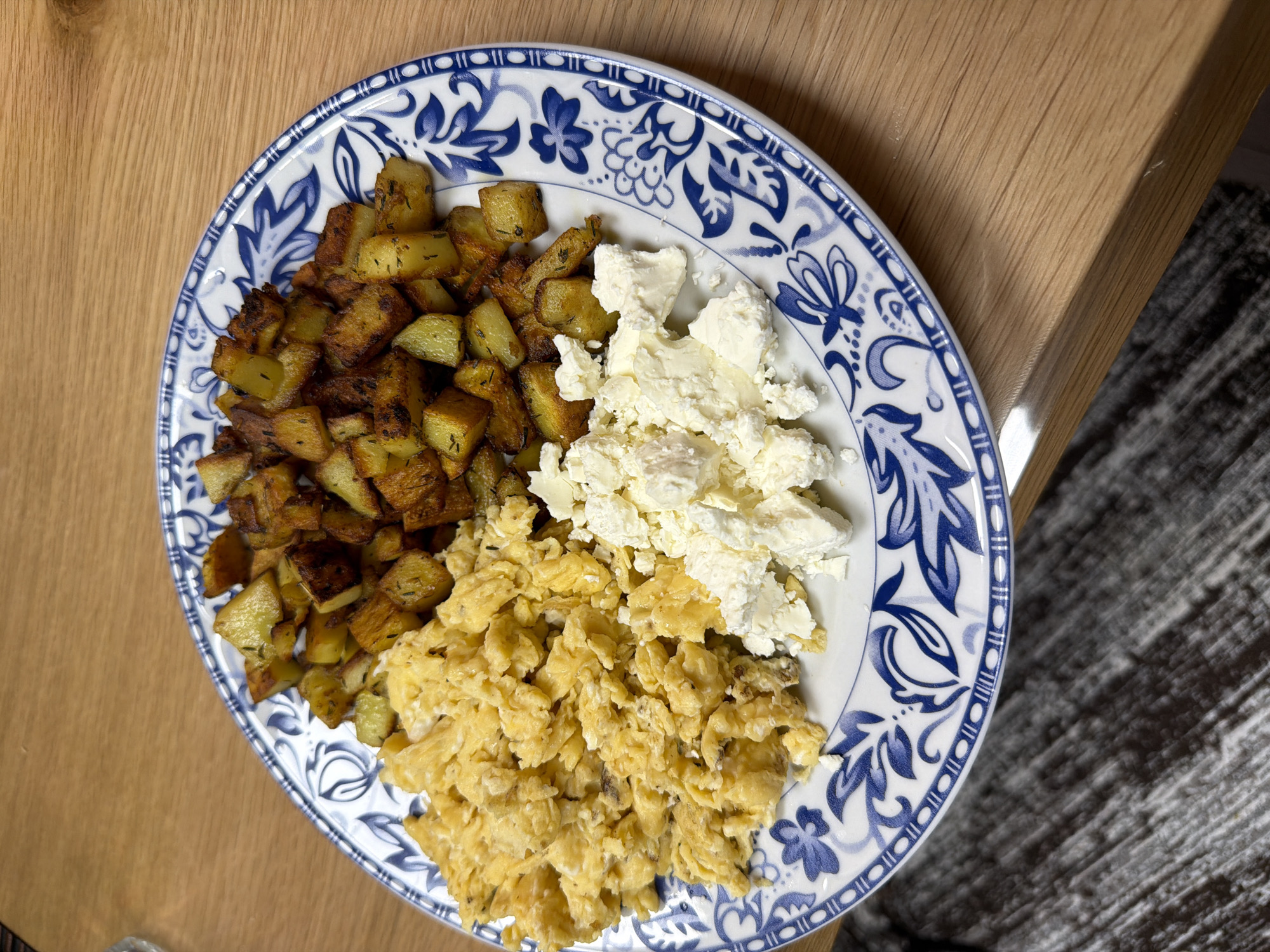 The image shows a plate of food, consisting of fried potatoes, scrambled eggs, and feta cheese, arranged in separate sections on a blue and white patterned plate. The plate rests on a wooden surface, which appears to be a table. Part of the surface is bare wood, and part is covered by a grey material. The scene does not provide any information that would place it in Panagyurishte, Bulgaria.
