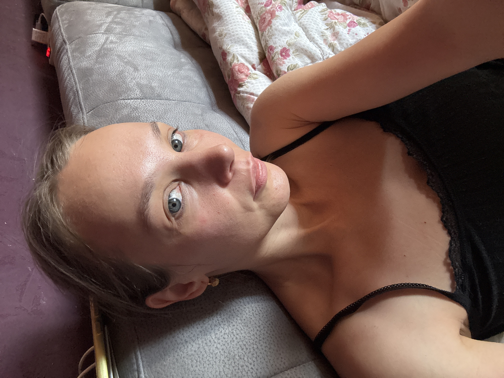 The image is a close-up, eye-level selfie of a young adult woman lying on a sofa. She is positioned on the left side of the frame, with her head tilted slightly towards the camera. She has light skin, light blonde hair pulled back, and blue-grey eyes. She is wearing a black tank top with lace trim. The background consists of a textured purple wall behind her head and a grey sofa. A floral-patterned blanket is visible on the right side of the frame. The overall lighting suggests an indoor setting, possibly a living room. The image does not contain information about the specific location in Überherrn, Germany.