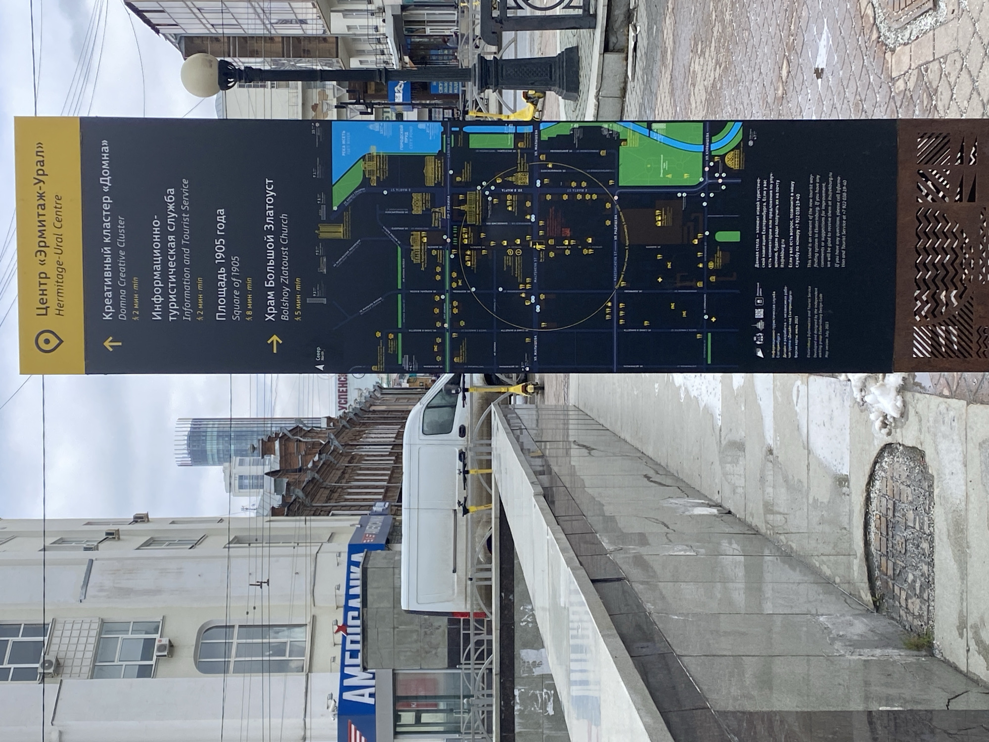Here is a detailed description of the image:
The image shows a street scene in Yekaterinburg, Russia, centered around a tourist information sign. The sign, predominantly black with yellow accents, displays a map of the city center and information about nearby attractions. It features the "Hermitage-Ural Centre," a creative cluster called "Domna," an information and tourist service, "Square of 1905," and the "Bolshoy Zlatoust Church." The sign also includes a QR code and contact information for the tourist service.
Behind the sign, there's a modern city street. The architecture visible consists of a mix of older buildings and a modern skyscraper in the background. A white delivery van is parked beside a gray walkway. Signs in Russian are present on buildings. The sky is overcast, suggesting cloudy weather.