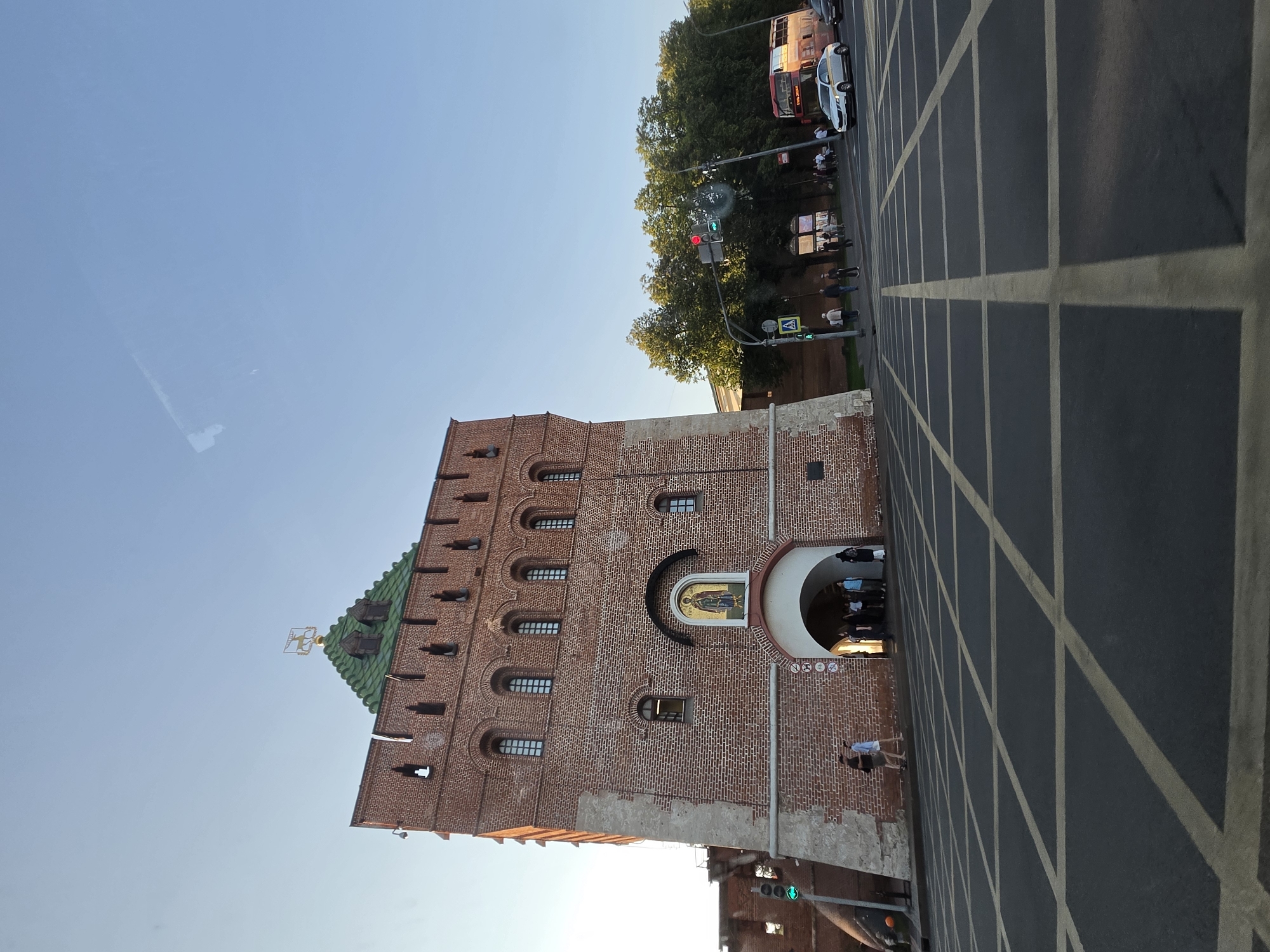 The image depicts the Dmitrovskaya Tower in Nizhny Novgorod, Russia, on a clear day with a bright blue sky. The tower, constructed of red brick, is a prominent feature of the scene, with its green roof and a gold emblem at its apex. An icon is visible above the arched entrance of the tower. Several people are present, with some walking near the tower and others further away, on the sidewalk. A double-decker bus and a white car are visible on the road, alongside a traffic light that shows a red and green signal. The foreground is dominated by a dark asphalt surface with a pattern of yellow lines, possibly indicating a pedestrian crossing or a parking area. Trees with lush green foliage line the street in the background.