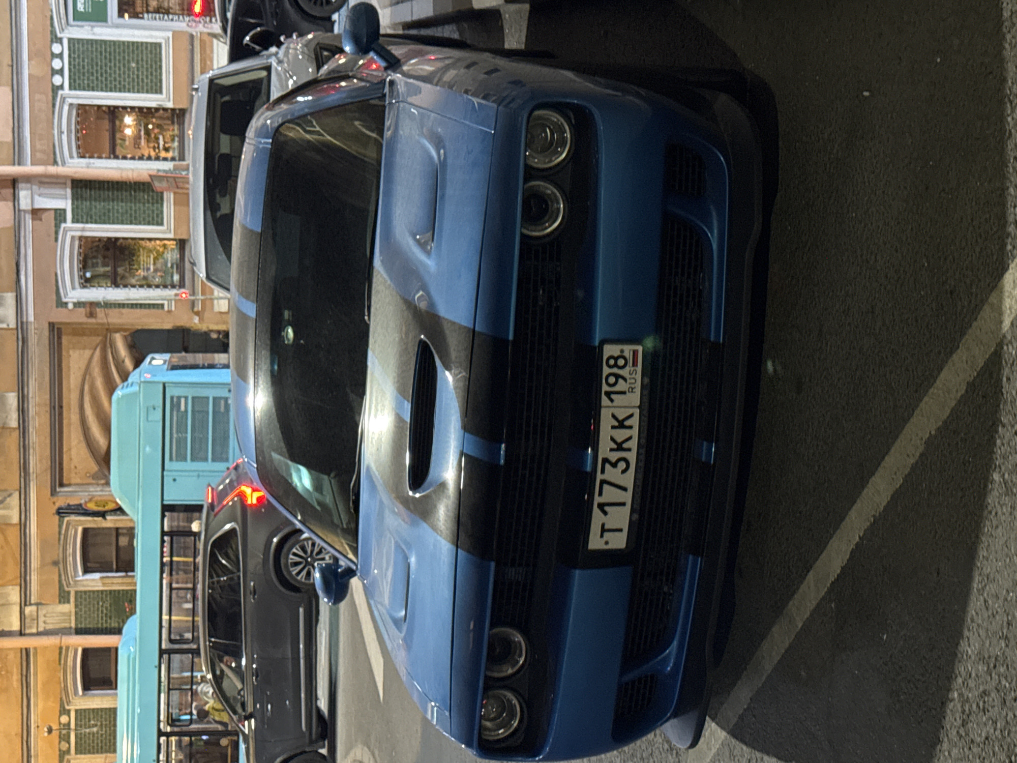 The image captures a blue Dodge Challenger muscle car parked on an urban street at night or dusk. The car, positioned prominently in the foreground, features a distinctive front grille, four circular headlights, and two black racing stripes running down its hood. A Russian license plate, "T 173KK 198 RUS", is clearly visible on the front bumper, with "198" being the region code for Saint Petersburg.
In the background, other vehicles are visible, including a dark grey SUV or sedan and a light blue double-decker tourist bus. The street is lined with multi-story buildings showcasing European architectural styles, characterized by arched windows and sections with green tiled facades. Lights emanate from within the buildings, reflecting in the windows. The ground is dark asphalt with a faint white line. No people are visible, and the scene depicts a static moment on a city street. The lighting suggests it is late evening or night, and the weather appears clear.