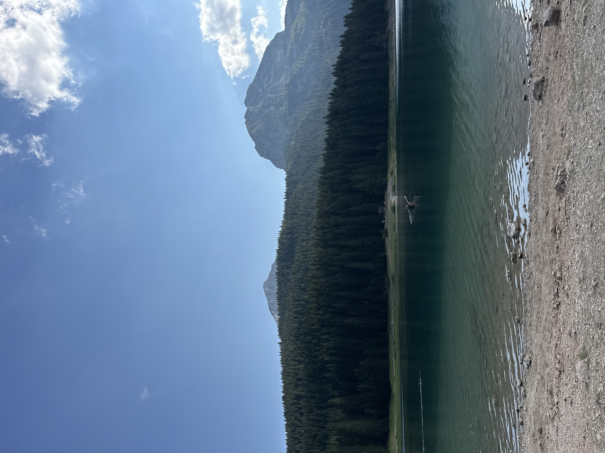 This is a vertical, eye-level, medium shot of a serene lake with a forested mountain range in the background, under a clear blue sky with scattered clouds. The foreground consists of a rocky shore leading into the calm, greenish-blue water. The water is clear enough near the shore to see the pebbles and stones beneath the surface, with rippling patterns reflecting the light.
In the middle ground, a dense, dark green coniferous forest covers the lower slopes of the mountains. Two distinct mountain peaks rise above the treeline. The left peak is smaller and cone-shaped, while the right peak is larger and more rugged, with visible rock faces and shadows. The sky above is a vivid blue, with wisps of white clouds scattered across the upper portion of the frame, catching the sunlight.
In the lake, a small rowboat with at least two occupants is visible in the middle distance, moving from left to right. Further to the left, near the shore, two small dark shapes that appear to be swimmers or waterfowl are present in the water. On the far left of the shore, a small wooden structure, possibly a shed or hut, can be seen. The overall scene suggests a peaceful, natural landscape. While the prompt mentions the city of Pitomine, Montenegro, the image itself does not contain any explicit elements that would identify it as such without external information.