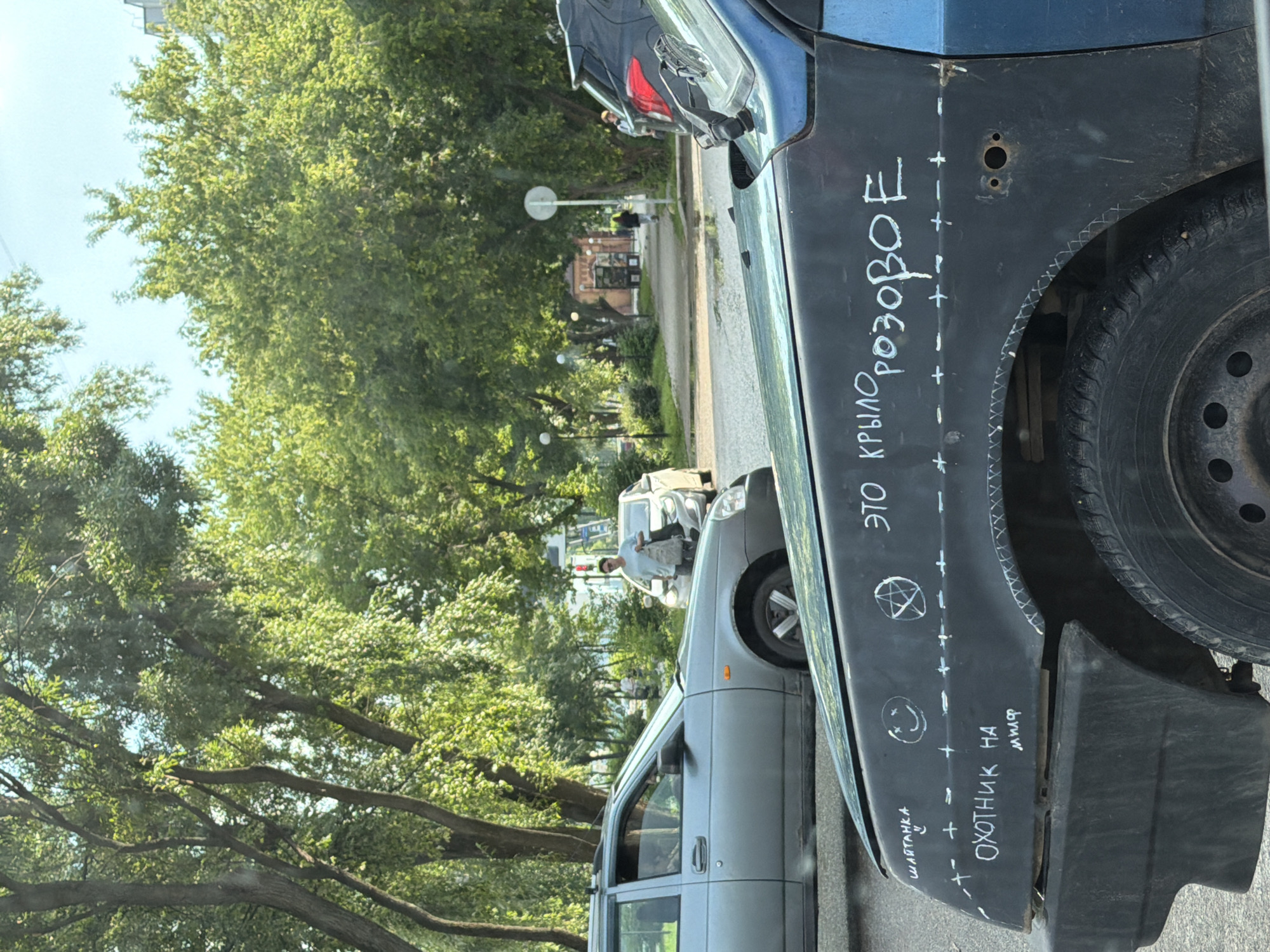 In Chaikovsky, Russia, on a sunny day, a light blue car is parked on a paved road, partially obscured by a dark blue car in the foreground. The dark blue car has white chalk writing on its hood and fender. The writing reads "ЭТО КРЫЛО РОЗОВОЕ" (This fender is pink) and also includes "ШАТАНКА" (Shaytan/demon), a smiley face, a pentagram, and the phrase "ОХОТНИК НА МИЛФ" (Hunter for MILF). White hash marks and crosses are drawn along a horizontal line on the fender. In the background, a silver car is visible, and a woman is walking away from it carrying bags. Trees line the street, and buildings are visible further in the distance. The sky is clear and blue.