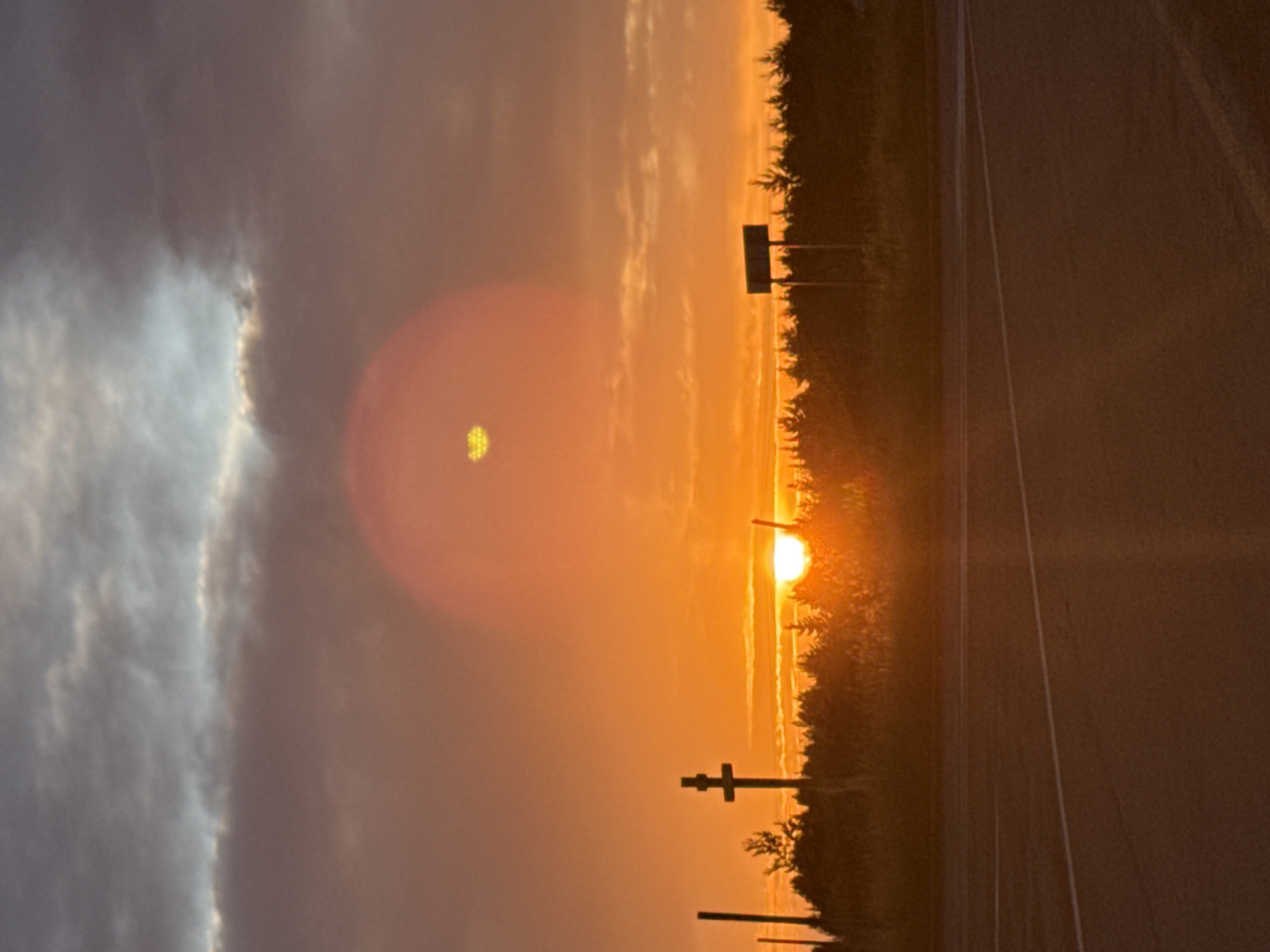 The image captures a sunset over a rural landscape, with a road in the foreground. The sky is filled with dramatic clouds, ranging from dark grey to vibrant orange and yellow, with the sun appearing as a bright orange disc near the horizon. A lens flare, a circular orange glow with a yellowish center, is visible in the upper portion of the sky.
In the middle ground, a silhouetted treeline stretches across the scene, with a few distinct objects protruding above it. To the left of the center, a dark, cross-shaped structure stands prominently. To its right, a tall, thin pole is visible. Further to the right, a rectangular sign mounted on two posts stands out against the treeline and sunset. The sun is partially obscured by the trees on the right side of the image.
The foreground consists of an asphalt road with white lane markings, suggesting it's a highway or a significant road. The overall impression is one of a remote, serene setting bathed in the warm light of a setting sun.
This scene is likely located in the vicinity of Гольяны, Russia, a village in the Tver Oblast, known for its rural character and natural landscapes, fitting the depiction of a sunset over a treeline and road. The presence of the cross structure might indicate a religious or memorial site, common in rural Russian landscapes.