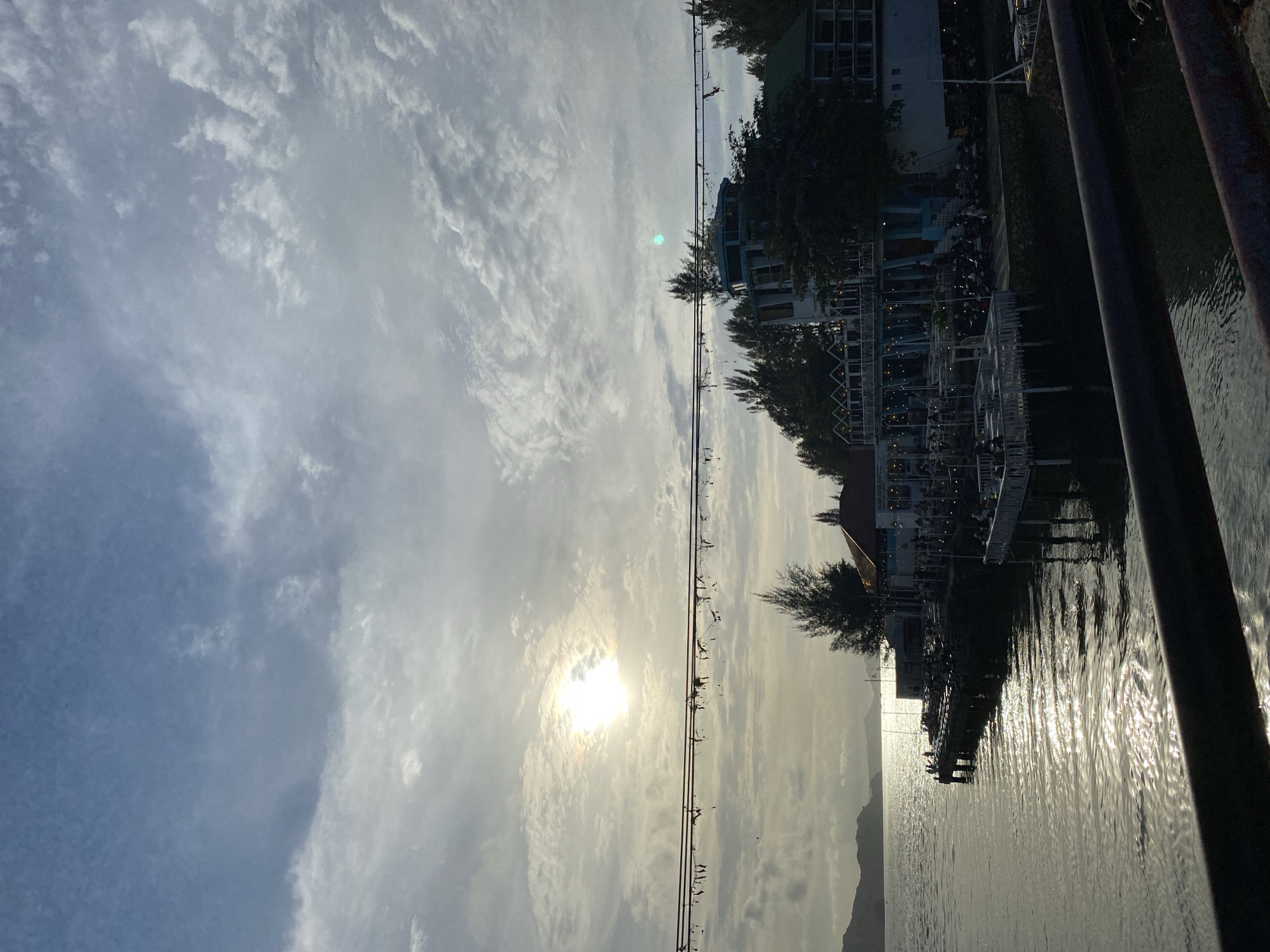 The image depicts a waterfront scene, likely in Banda Aceh, Indonesia, during what appears to be late afternoon or early evening. The sun, partially obscured by clouds, casts a bright, shimmering reflection on the calm water. In the foreground, a dark railing or barrier runs horizontally across the bottom of the frame.
The middle ground features a pier or boardwalk extending into the water, lined with tables and chairs, suggesting an outdoor dining or seating area. Several people are visible on the pier, some seated at tables, indicating a relaxed atmosphere. A multi-story building with white and teal accents stands on the waterfront, partially covered by trees. This building has multiple levels of balconies and an unusual, rounded top section. Various vehicles, likely motorcycles, are parked along the waterfront in front of the building. The architecture suggests a commercial establishment, possibly a restaurant or cafe.
The background shows a body of water leading to a distant shoreline with hills or mountains. The sky is filled with a mix of clouds, with lighter patches indicating the sun's position. Overhead, a series of wires or cables with what appear to be small, tattered decorations or debris strung between them traverse the upper portion of the image. The overall impression is one of a scenic, possibly popular, waterfront location.
