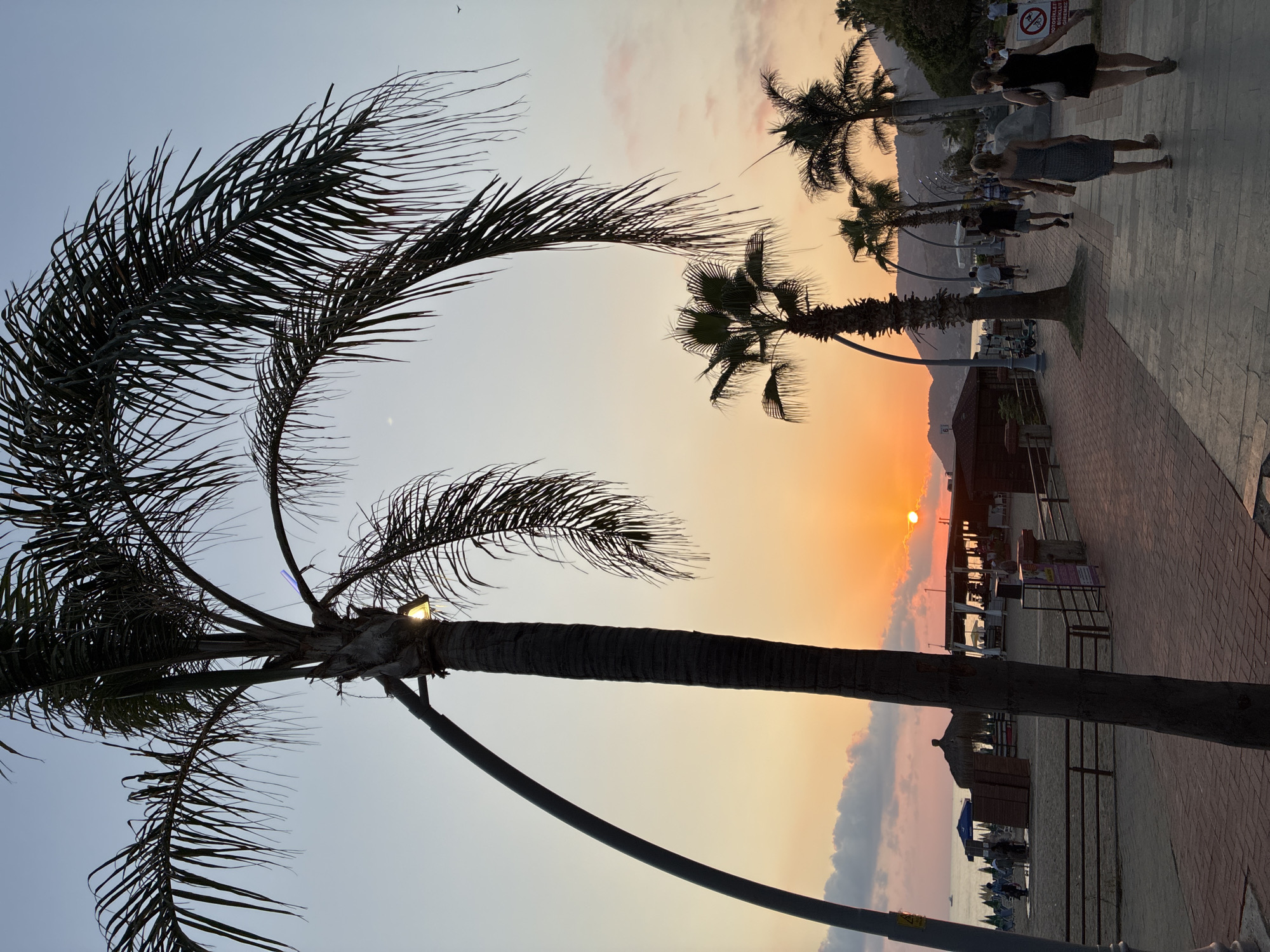 This image depicts a sunset scene along the Alanya promenade in Turkey. The sky displays a gradient of pale blue, orange, and pink hues. Several palm trees are scattered throughout the scene, with their fronds silhouetted against the sky. In the foreground, a paved walkway is visible, with people strolling along it. To the left, a beach area with umbrellas and some structures suggestive of beachside cafes or shops is present. The sun, a bright orange orb, is low on the horizon, partially obscured by clouds. The overall atmosphere is tranquil and evening-oriented.