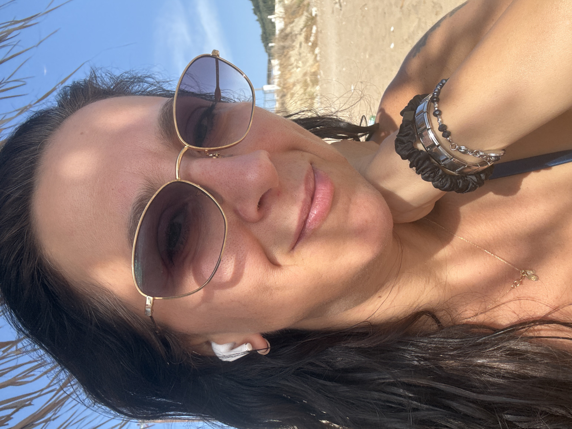 In Agios Georgios, Greece, a woman with dark hair and sunglasses is taking a selfie on a sandy beach. She is wearing white wireless earbuds and a thin gold necklace with a small charm. She has several bracelets on her left wrist, including a black scrunchie bracelet and a silver charm bracelet. The background shows a blue sky with some clouds and a sandy embankment with sparse vegetation. A palm frond from a beach umbrella is visible in the upper left corner of the image.