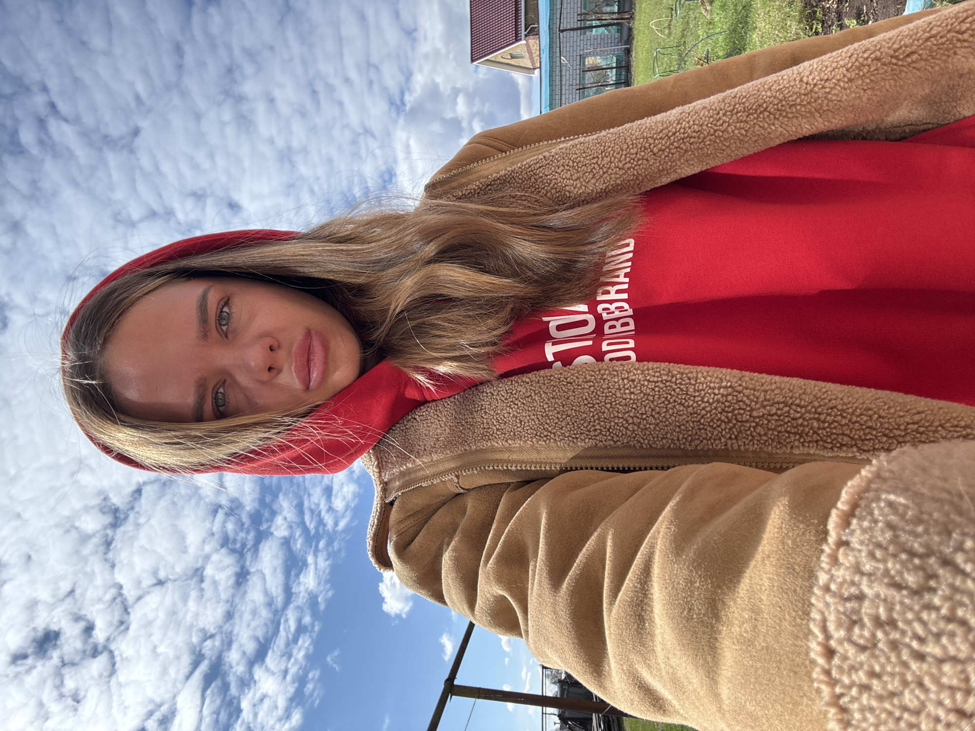 The image is a tilted, close-up selfie of a young woman taken outdoors, primarily featuring her face and upper body against a vast blue sky. The perspective is from a low angle looking upwards, creating an unusual composition where her head is at the bottom of the frame and the sky dominates the top.
The subject is a woman with long, light brown hair flowing over her shoulders. She has green eyes and full lips, looking directly at the camera with a neutral expression. She is wearing a vibrant red hooded sweatshirt, with the hood partially framing her face. Over the hoodie, she wears a warm-looking tan or light brown jacket or vest, prominently featuring a textured, fuzzy faux shearling lining visible around the collar and sleeves.
The background consists mainly of a bright blue sky filled with numerous small, scattered white clouds, indicative of a clear and sunny day. In the upper portion of the frame, behind the woman, elements of a rural or suburban environment are visible: green grass, the dark red tiled roof of a building or house, and a dark fence-like structure.
Visible white text on the red hoodie is partially cut off but shows "TO" and "DIBBRAND," likely part of a brand name such as "ODDBRAND" or "GOODBRAND." The overall setting suggests a pleasant outdoor environment, possibly a garden or backyard, consistent with the location context of Bol'shoye Kozino, Russia.