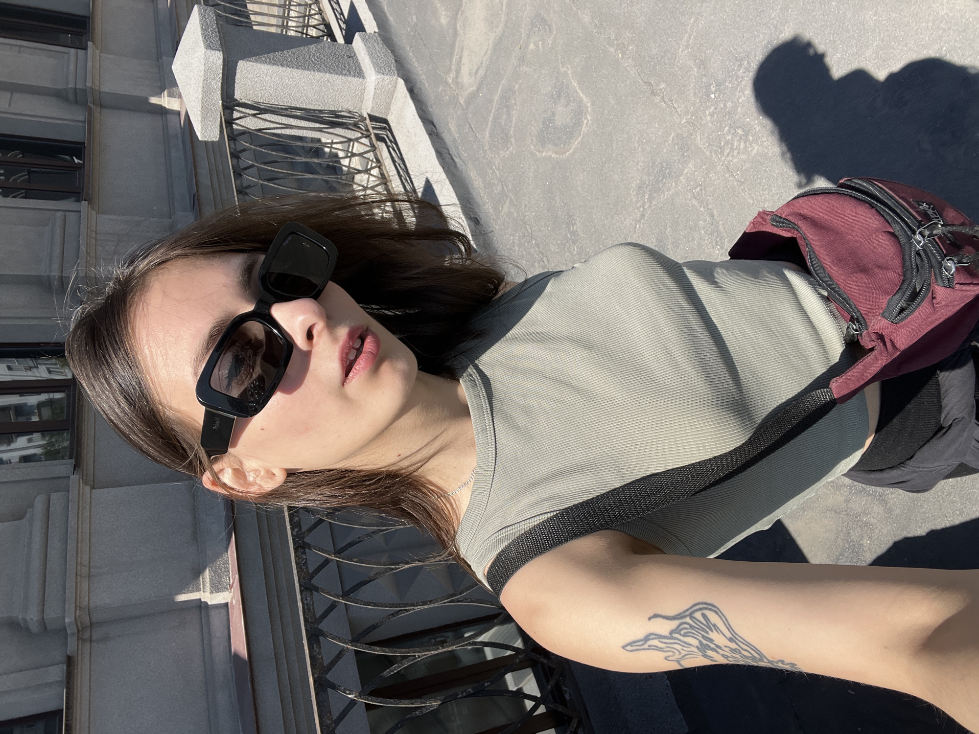 A young person is captured in a sunlit outdoor selfie, likely in Krasnoyarsk, Russia. The subject is shown from the chest up, with their head slightly tilted, looking directly towards the camera. They wear black, square-framed sunglasses, with "Polaroid" visible on the right arm. Their lips are slightly parted, with a hint of light pink lip color. Long, dark brown hair frames their face and falls over their shoulders.
The person is dressed in a light olive green ribbed short-sleeved top and wears a dark red or burgundy sling bag with multiple black zippers, draped across their body. A black strap for the bag is visible over their left shoulder, and a thin silver necklace chain adorns their neck. A distinct black-outlined tattoo, featuring a fluid, abstract, or wave-like design, is prominent on their right forearm.
The background consists of a tall building with a grey, possibly stone or concrete, facade featuring architectural details and dark windowpanes. In front of the building, a decorative metal railing or fence is visible. The foreground and right side of the image show light grey pavement illuminated by strong sunlight, casting sharp shadows, including a large shadow that resembles a person's upper body and head on the pavement to the right, suggesting the photographer's own shadow. The bright light and harsh shadows indicate a clear, sunny day, likely around midday.
