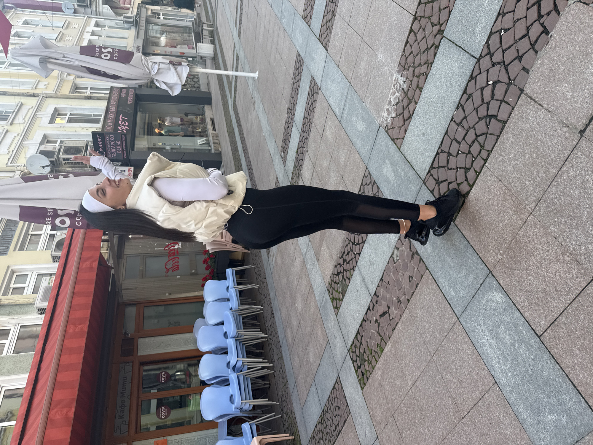 The image shows a woman standing on a cobblestone and concrete sidewalk in Gabrovo, Bulgaria. She is facing the camera's left and looking upwards, possibly at a building or sign, with a smile. She is wearing a white headband, a white long-sleeved shirt, a cream-colored puffer vest, and black leggings. The woman's hair is long and dark. She is also wearing black sneakers.
Behind her are a building with a red awning, tables, and chairs. A Costa Coffee shop is visible on the building's front. To the left, there are buildings with white umbrellas over the tables. Signs are present in Cyrillic script. The sidewalk is made of gray and beige concrete pavers and cobblestones. The building on the left appears to be an apparel store with mannequins in the display window.