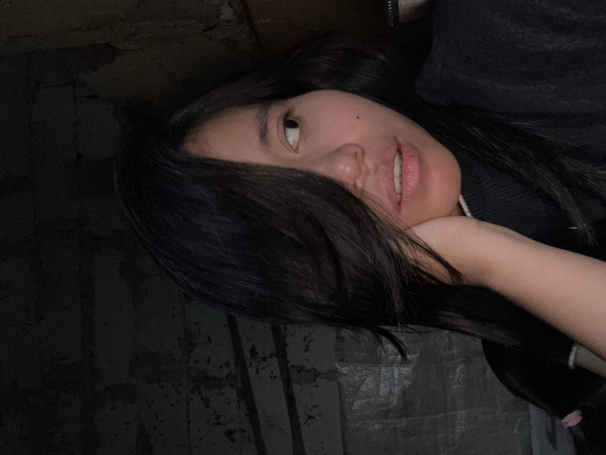 In a dimly lit indoor setting, a young woman with dark hair, partially covering her face, is captured in a close-up portrait. She is wearing a black turtleneck. Her left hand rests on her chin and cheek, with her fingers gently touching her face. Her gaze is directed to her right, with her mouth slightly open. The background appears to be a textured, dark wall, possibly brick, with some horizontal lines suggesting construction or shelving. To the left of the woman, a plastic-covered surface is visible. The overall lighting is low, creating shadows and highlighting the contours of her face. The specific location of East Jakarta, Indonesia is not visually discernible from the provided image.
