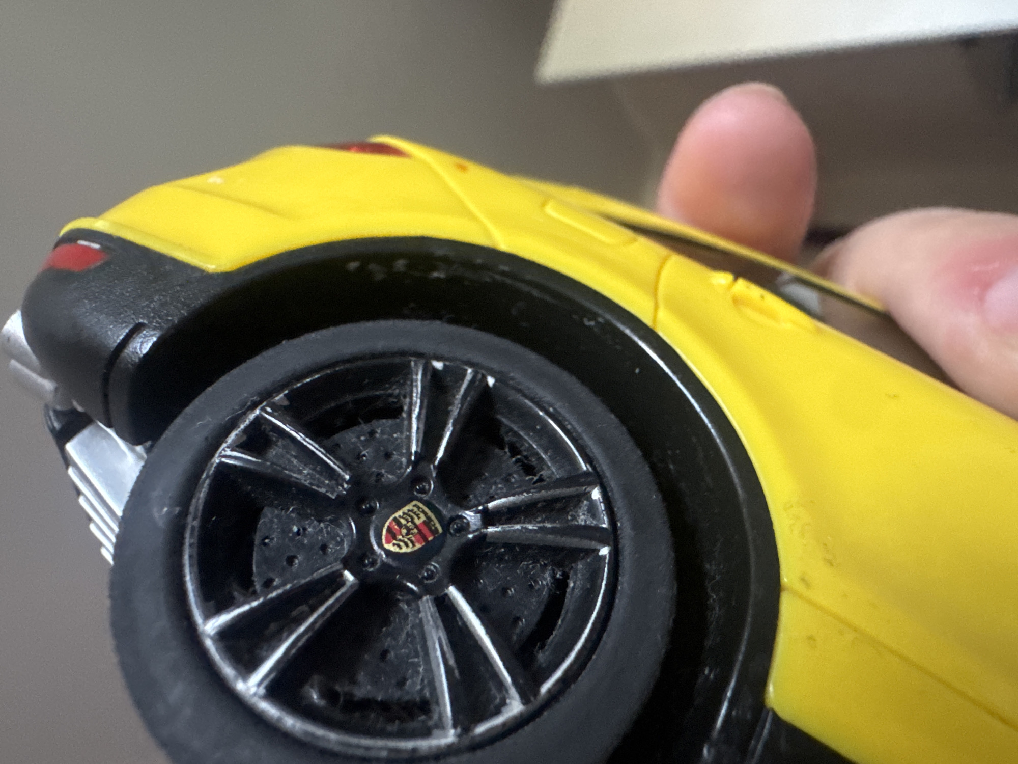 The image shows a close-up, inverted view of a yellow toy car, likely a Porsche, held by a person's fingers. The car is upside down, with its black tire and detailed silver wheel rim prominently visible. The wheel has a central emblem, which appears to be a Porsche logo. The body of the car is bright yellow, and there are some dark accents, possibly from the car's interior or trim. Dust or debris is visible on the wheel and the surrounding car body. The background is out of focus, appearing as a muted, neutral color, likely a wall or surface. The image does not contain any contextual information that would allow for its location to be identified as Asha, Russia.