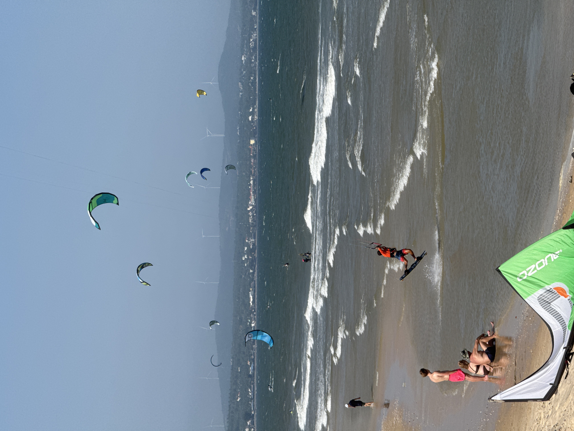 The image depicts a busy beach scene in Phan Thiet, Vietnam, likely Mui Ne, a popular kiteboarding destination. The ocean is active with moderate waves, and the sky is clear with a slight haze. Multiple kiteboarders are visible on the water, with some performing jumps and tricks. The kites, in various colors including blue, green, yellow, and multicolor, are scattered across the sky. In the foreground, a green and white kite is laid out on the sand. Several people are on the beach; some are standing and observing the kiteboarders, while others are sitting or relaxing on the shore. A few individuals are wading in the shallow water. In the background, a coastline with buildings and rolling hills dotted with wind turbines can be seen. The overall atmosphere suggests a lively day for watersports and beach activities.