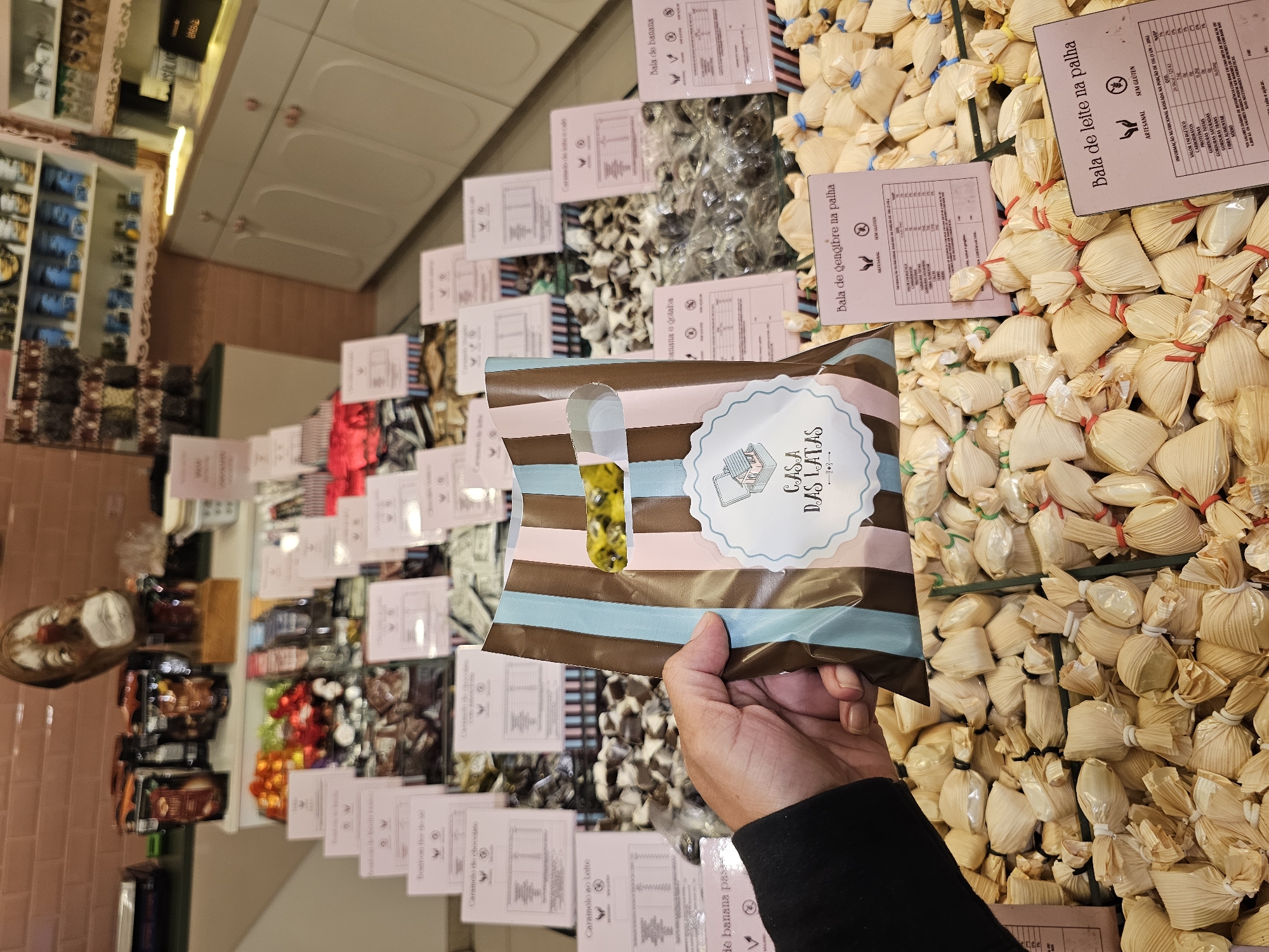 A hand holds a striped bag labeled "Casa Das Latas" in a candy store displaying various sweets. The shop sells items such as caramelos de chocolate, caramelos ao leite, balas de banana, balas de gengibre na palha, and balas de leite na palha. The location of this store is likely in Armação dos Búzios, Brazil.