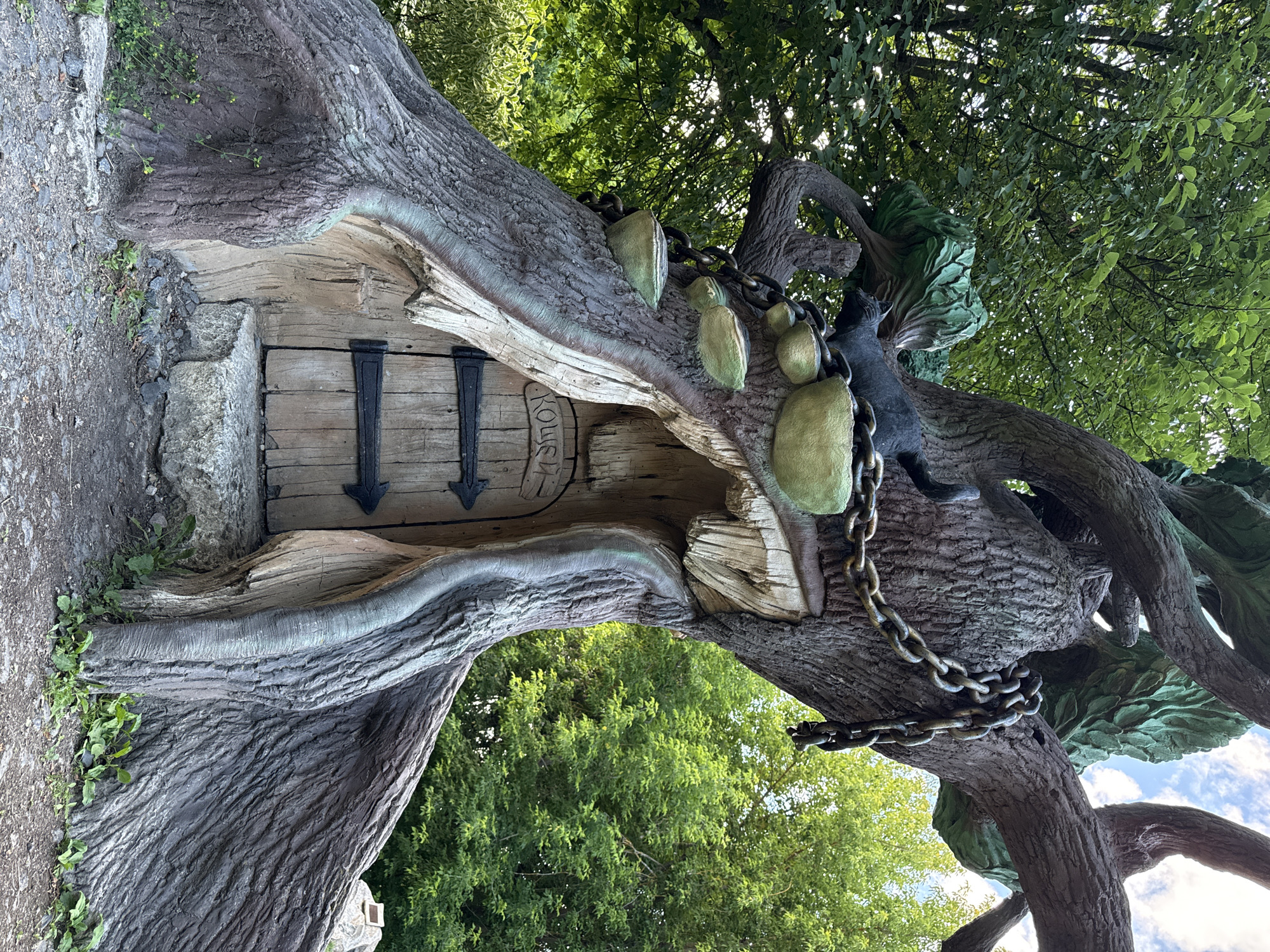 This image captures a whimsical, artfully sculpted tree with a miniature door and whimsical elements, located in the city of Letniy Otdykh, Russia. The tree, appearing to be a man-made artistic creation rather than a natural growth, features a textured, bark-like exterior painted in shades of brown and grey with subtle green accents. Carved into its trunk is a small, rustic wooden door, complete with black metal hinges and decorative arrow-shaped handles. Above the door, the word "Кощей" (Koschei) is inscribed in stylized letters.
The tree is adorned with several large, round, bowl-like protrusions painted in shades of green and yellowish-green. A black, stylized cat figure is perched on one of the branches, gazing downwards. A thick, dark metal chain wraps around the trunk and branches, adding to the fantastical theme. The sculpture is set against a backdrop of lush green foliage and trees, with a partly cloudy sky visible in the upper right corner. The ground around the base of the tree is covered in a mixture of gravel, dirt, and some scattered stones, with small green plants growing in the crevices. The overall impression is that of an enchanted, storybook-like setting within a park or recreational area.