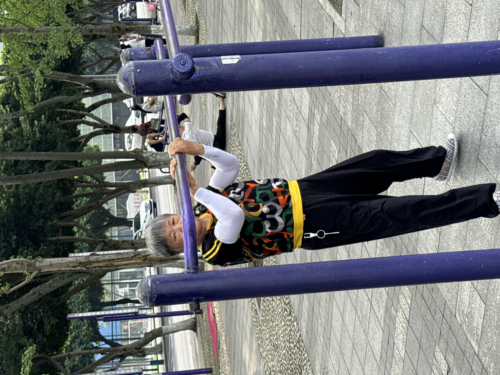 This image, captured in Jiangjin, China, depicts an outdoor public exercise area. In the foreground, an older East Asian woman with short, grey hair is performing an exercise on a purple metal horizontal bar. She wears a black short-sleeved top with a vibrant, abstract pattern in green, orange, white, and black, featuring a yellow collar and hem. Long white sleeves are worn underneath. She is dressed in black loose-fitting pants and patterned slip-on shoes. A silver keyring with a key is clipped to her right hip. She grasps the bar with both hands, palms facing up, and has a faint smile.
Behind her, another person lies on a red mat on the paved ground to the left, stretching. Further back on the right, another individual sits on a similar purple exercise apparatus, also stretching, while a third person stands nearby. The ground is paved with patterned grey and light grey rectangular tiles. Numerous green-leafed trees, some with white-painted trunks, line the area, creating a park-like environment. A road is visible beyond the trees, with several vehicles, including a white sedan. A white sign with the Chinese characters "体育公园" (Sports Park) is visible in the distant background, indicating the public recreational nature of the location.