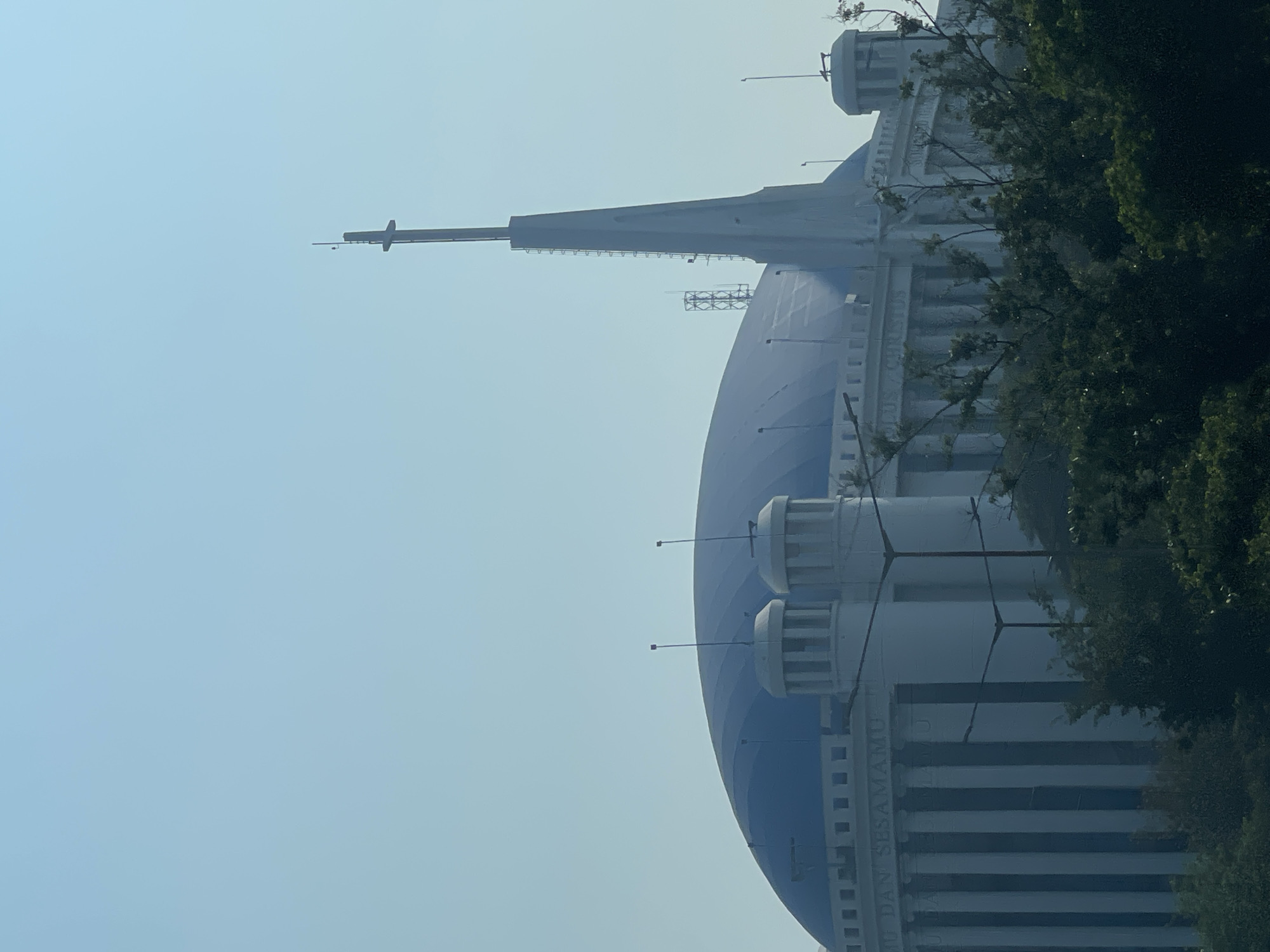 A large, modern building with a prominent blue domed roof dominates the frame, set against a clear, pale blue sky. The building features a distinctive tall, spire-like structure on its right side, topped with a cross. The facade of the building is characterized by white columns and what appears to be lettering. The foreground is filled with dark green foliage, obscuring the lower portion of the structure. The overall scene suggests a bright, possibly sunny day in an urban environment. The building is the Immanuel Church in Central Jakarta, Indonesia.