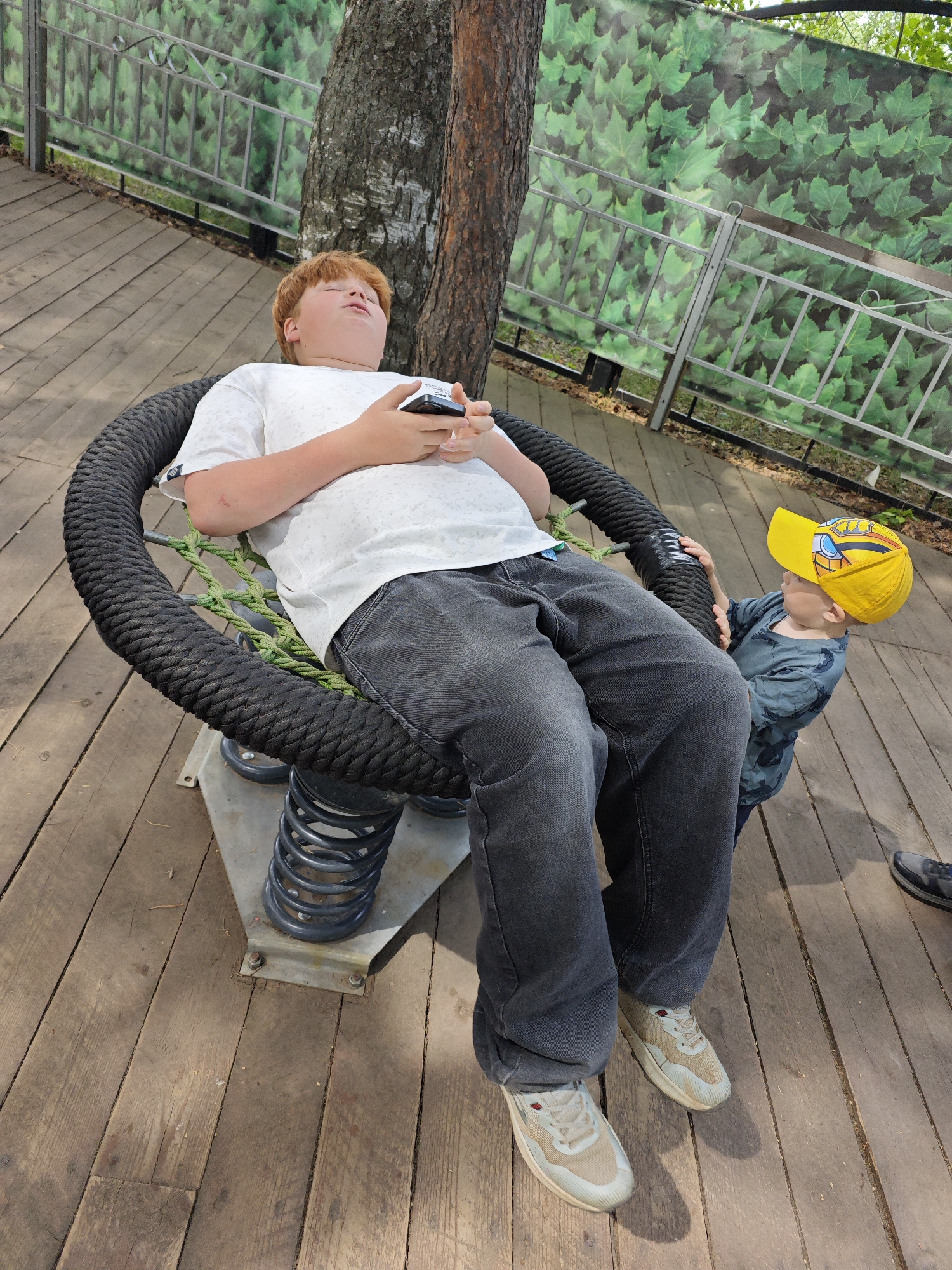 The image shows a young red-haired boy lying back in a black rope-covered circular chair, holding a device in his hands. The chair has a metal base with springs. A smaller child wearing a yellow baseball cap stands next to the chair, holding onto the rope. The chair is on a wooden deck near a tree trunk. A fence with green leafy patterned fabric behind it is visible. The image was taken in the city of Chaikovsky, Russia.
