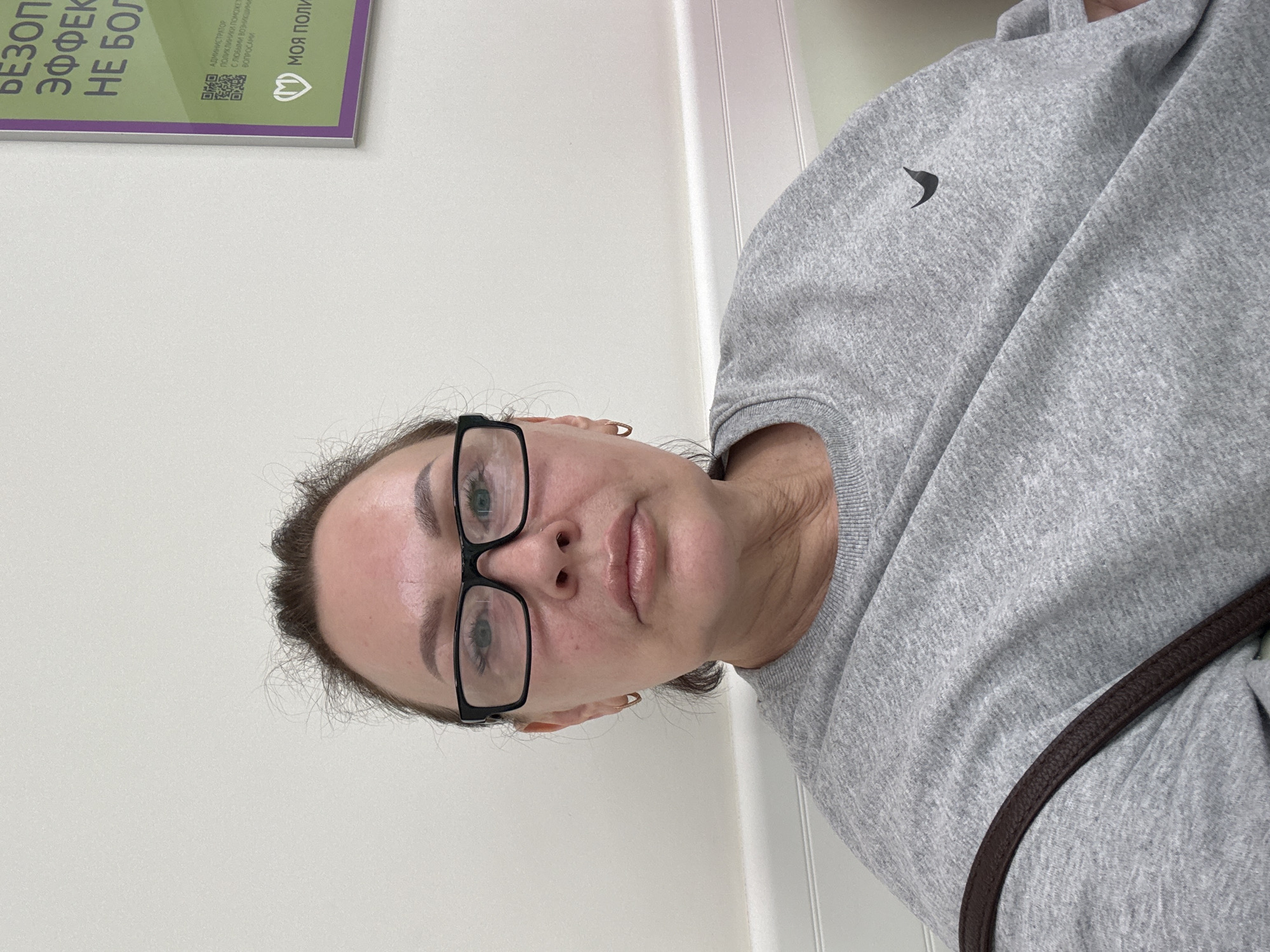 This is a selfie of a woman wearing glasses and a grey sweatshirt. She has light skin and her hair is pulled back. The background appears to be indoors, with a white wall and a light-colored door or partition. On the left side of the image, a sign with Russian text is partially visible. The text on the sign includes "БЕЗОП", "ЭФФЕК", and "НЕ БОЛ", which are likely abbreviations for words related to safety, effectiveness, and pain. Below this, there is a QR code and more text, including "АДМИНИСТРАТОР", "ПОЛИКЛИНИКИ ПОМОЖЕТ", and "МОЯ ПОЛИ". The woman's expression is neutral. The lighting suggests it is daytime. The overall setting appears to be a waiting area or a public building, possibly a clinic or medical facility given the text on the sign.