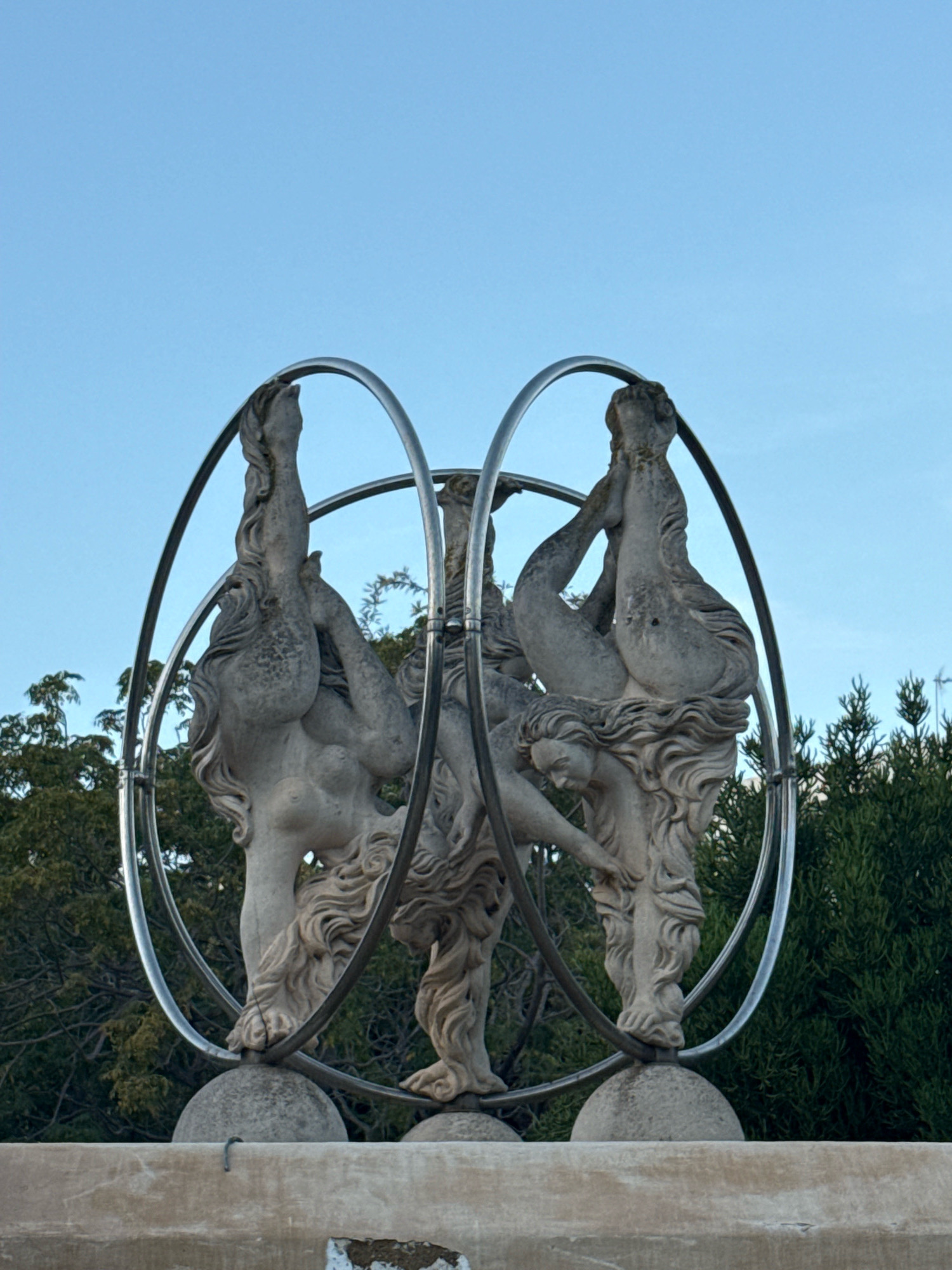 An outdoor stone sculpture of three female figures intertwined within two chrome rings sits on a textured concrete pedestal. The figures are rendered in a classical style, with flowing hair and draped bodies, some appearing to be in acrobatic poses. The sculpture is set against a clear blue sky and a backdrop of dense green trees. The bottom portion of the image shows a rough, weathered stone wall or base. The scene appears to be in a garden or public space. This sculpture is located in the Dalt Vila, the old town of Ibiza, Spain.