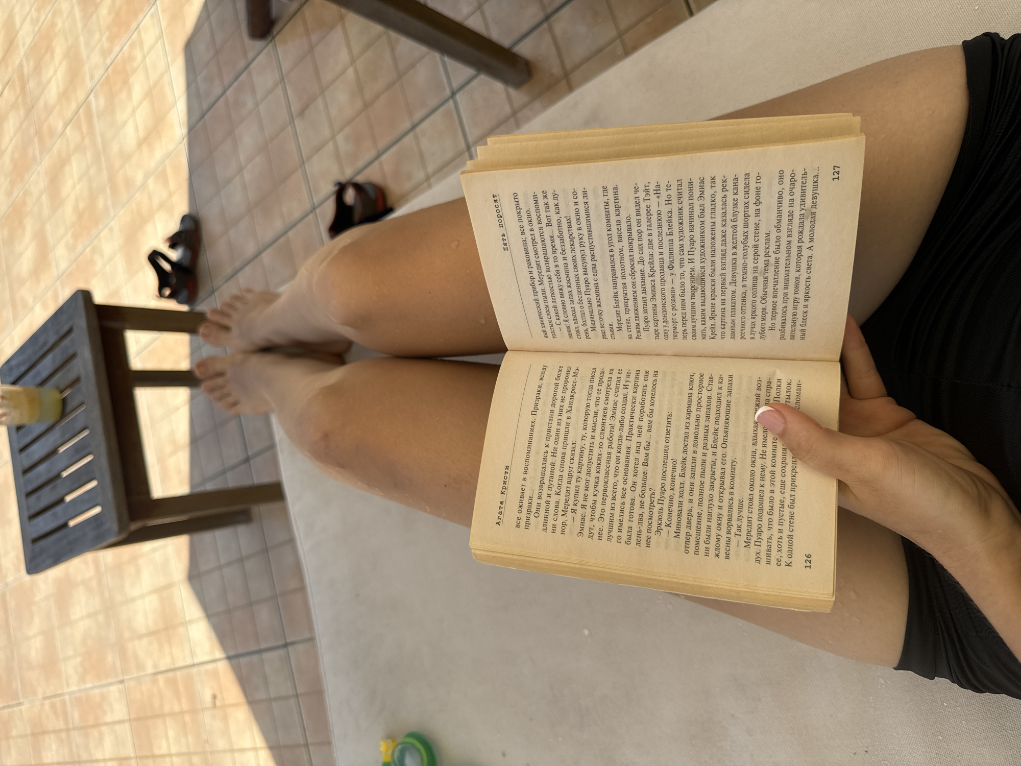 A person sits with their legs extended, holding open a book titled "Five Little Pigs" by Agatha Christie. The book is in Russian. The person is wearing a black garment. A stool sits on the tan, tiled floor, next to a pair of dark sandals. A drink with a straw rests on the stool. The background shows a tan-tiled surface, suggesting an outdoor area or patio. The photograph appears to be taken from an elevated perspective, looking down at the person and the surrounding objects. The location in Phu Thuy Ward, Vietnam, cannot be confirmed or inferred based on the image.