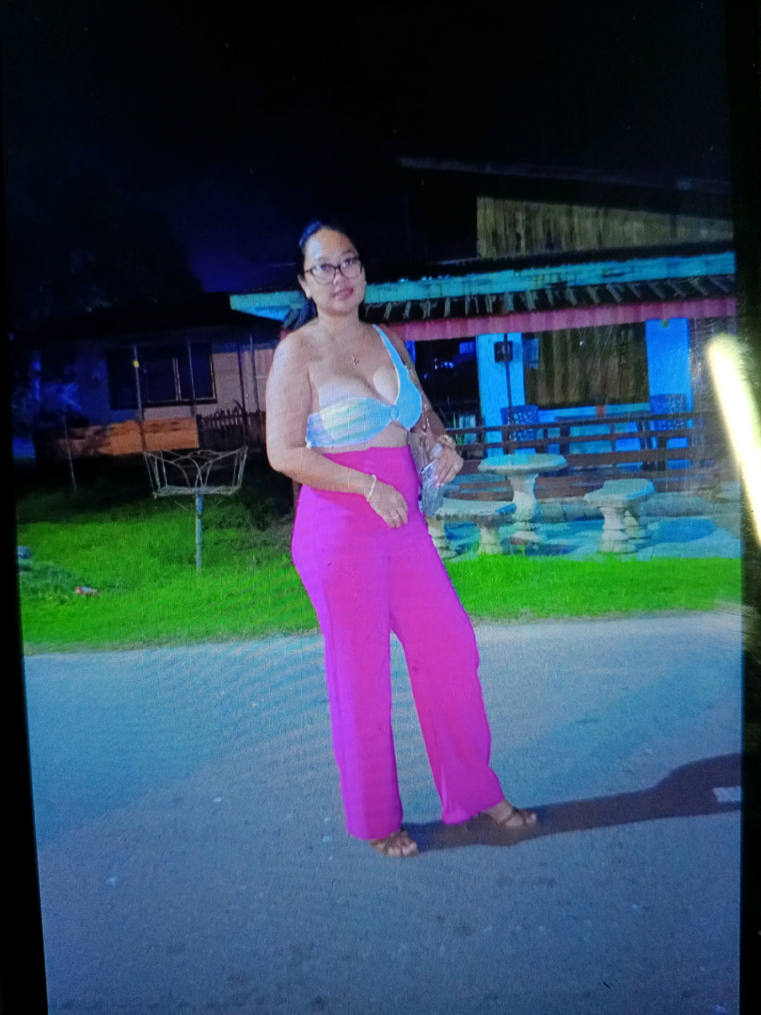 A person wearing a blue and white strapless top and bright pink wide-leg pants stands on a paved surface at night. They are also wearing glasses and sandals. Behind them, there is a grassy area with some outdoor seating structures and a building with lights. The location appears to be Noord, Suriname.