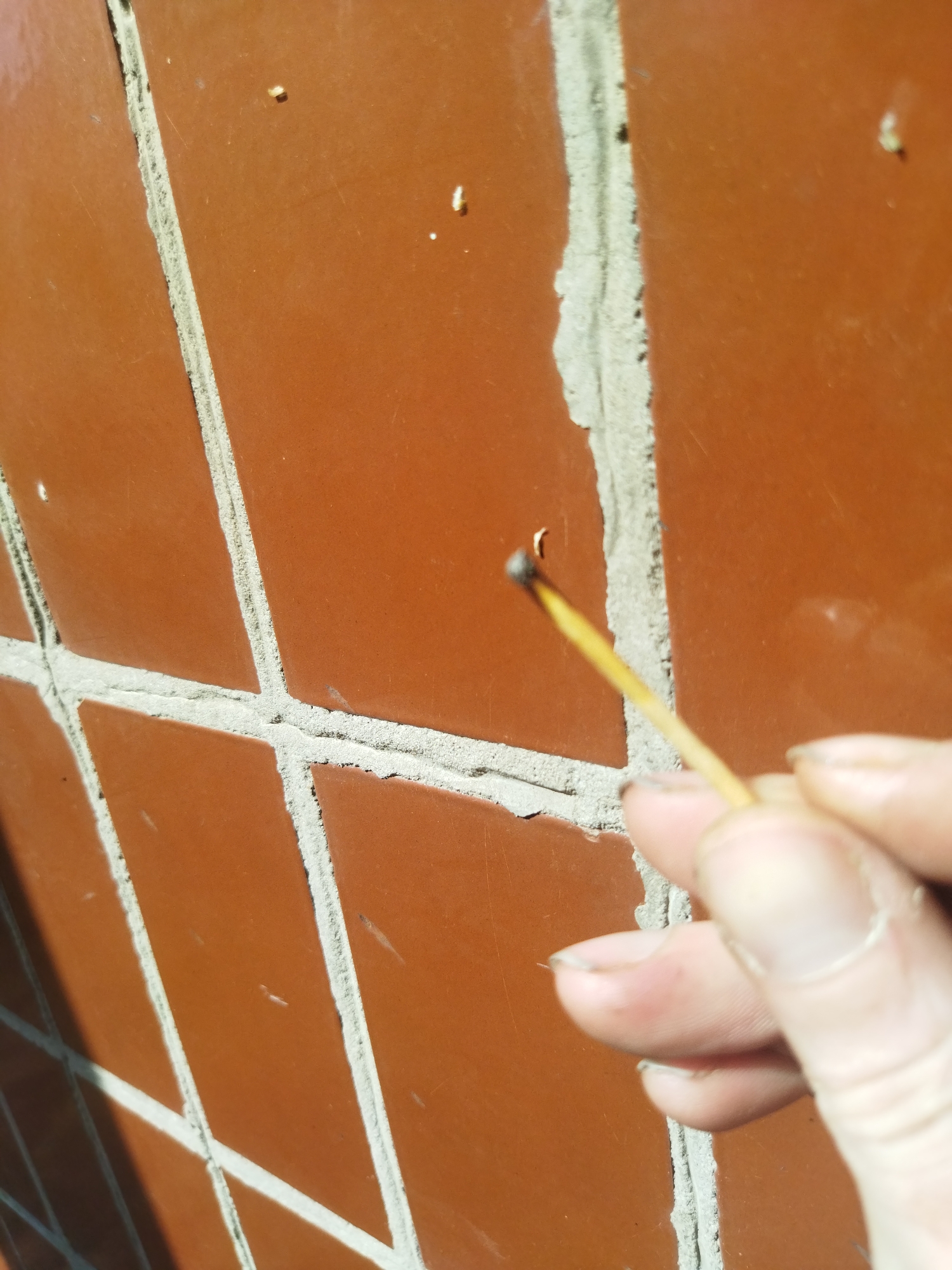 A hand holds a burnt match stick in front of a tiled wall. The wall is composed of orange tiles with gray grout. The location is not discernible from the provided image. No other people or subjects are visible. The only action is the holding of the match.