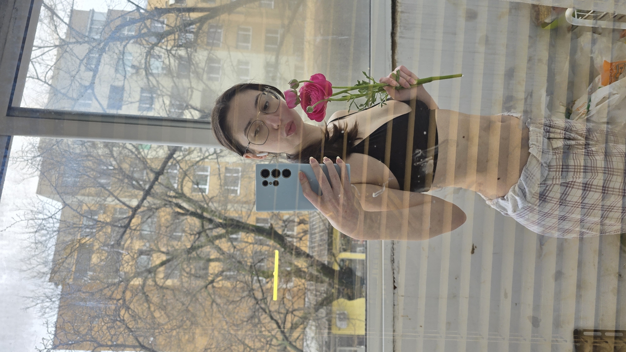 A woman in a black bra and plaid shorts is taking a selfie in a mirror with a light blue smartphone. She is holding pink flowers in her hand. The reflection in the mirror shows a building and trees outside. There is no indication of the location as Stara Zagora, Bulgaria in the image.