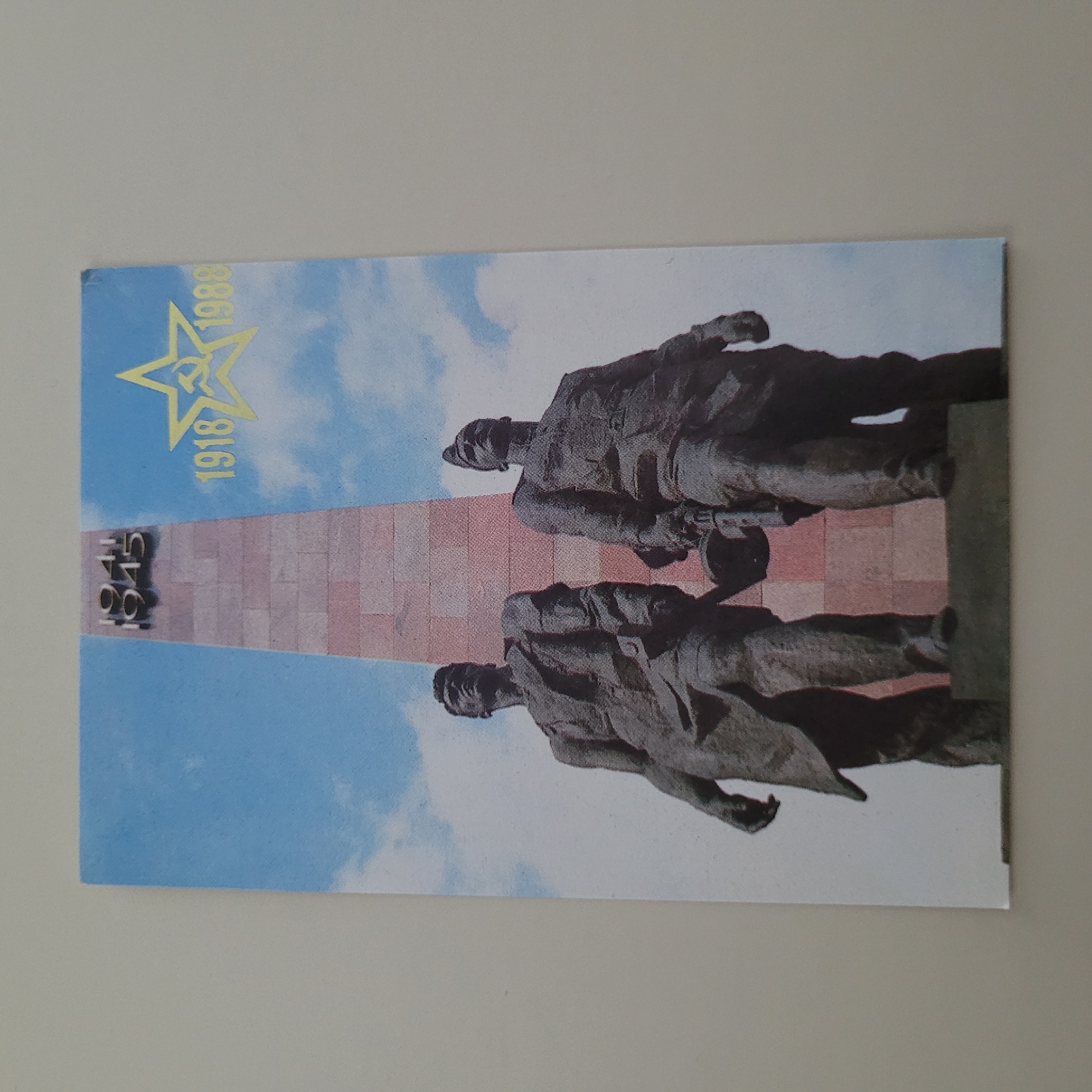 The image shows a postcard depicting a monument with two soldier statues in the foreground and a tall obelisk behind them. The obelisk has the numbers "1941" and "1945" inscribed on it. To the right of the obelisk, a yellow star with a hammer and sickle is prominently displayed, along with the numbers "1918" and "1988". The sky in the background is blue with scattered white clouds. The postcard is laid flat on a neutral-colored surface. The monument is the Monument to the Liberators of Riga from the German Fascist Invaders in Jūrmala, Latvia.
