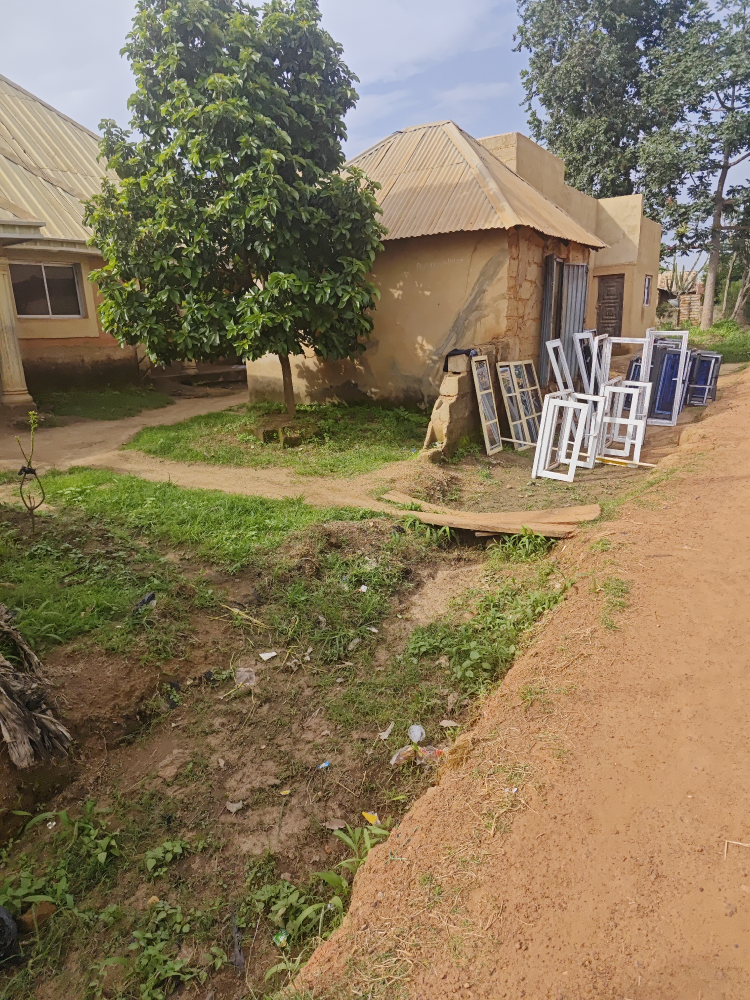The image depicts an outdoor scene in Bukuru, Nigeria, likely a residential or commercial area under construction or renovation. A traditional-style building with a corrugated metal roof is prominent, situated beside another building with a similar roof and a pale yellow exterior.
In front of the buildings, a collection of new window frames, primarily white and some with blue panes, are stacked neatly. They are leaning against the wall of the building or propped up on the ground. Several of these frames are of varying sizes and designs, suggesting they are intended for different parts of the structure.
To the left of the buildings, a mature tree with dense green foliage provides shade, its branches extending over the scene. The ground is a mix of dirt paths, patches of green grass, and areas of mud, with some scattered debris visible in the foreground. A small wooden plank crosses a ditch or channel in the grass.
The overall atmosphere is one of everyday activity, possibly related to construction or preparation for new installations. The lighting suggests it is daytime, with bright sunlight and a partly cloudy sky. The setting appears to be in a rural or semi-urban area of Nigeria, given the architectural style and the natural vegetation. No people are visible in the frame, and there is no discernible text in the image.
