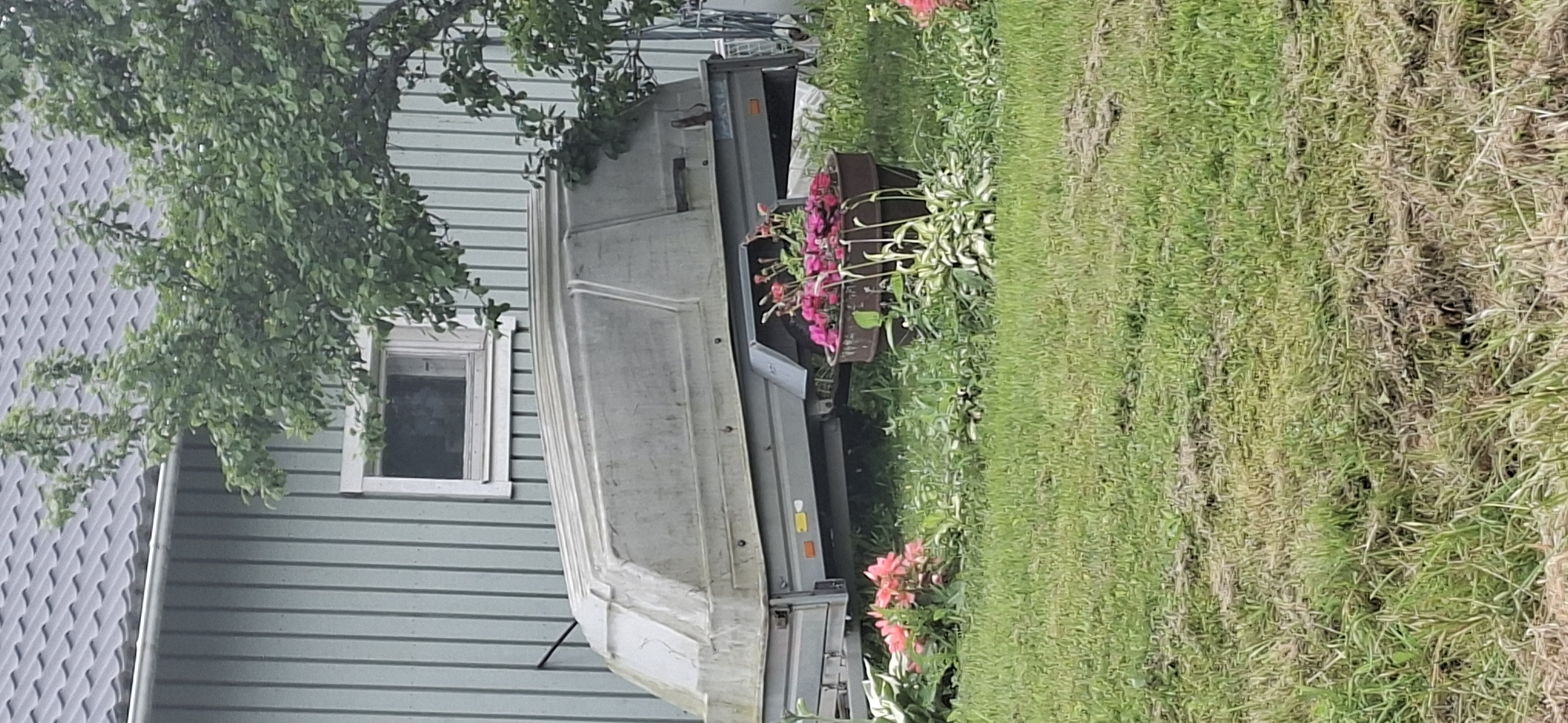 A light gray colored trailer with a domed cover is parked in front of a light green wooden house. The trailer is positioned horizontally in the frame, with its front to the left and back to the right. The house has vertical siding and a dark gray shingled roof. A single window with a white frame is visible on the house wall behind the trailer. A large tree with green leaves is situated to the right of the trailer, partially obscuring the house and the trailer's right side. In the foreground, a patch of green grass is visible, with some areas appearing dry and matted. A large planter filled with pink flowers sits on the grass in front of the trailer. Another cluster of pink flowers is seen to the left of the planter, in front of the trailer. The scene appears to be outdoors, likely in a residential area.