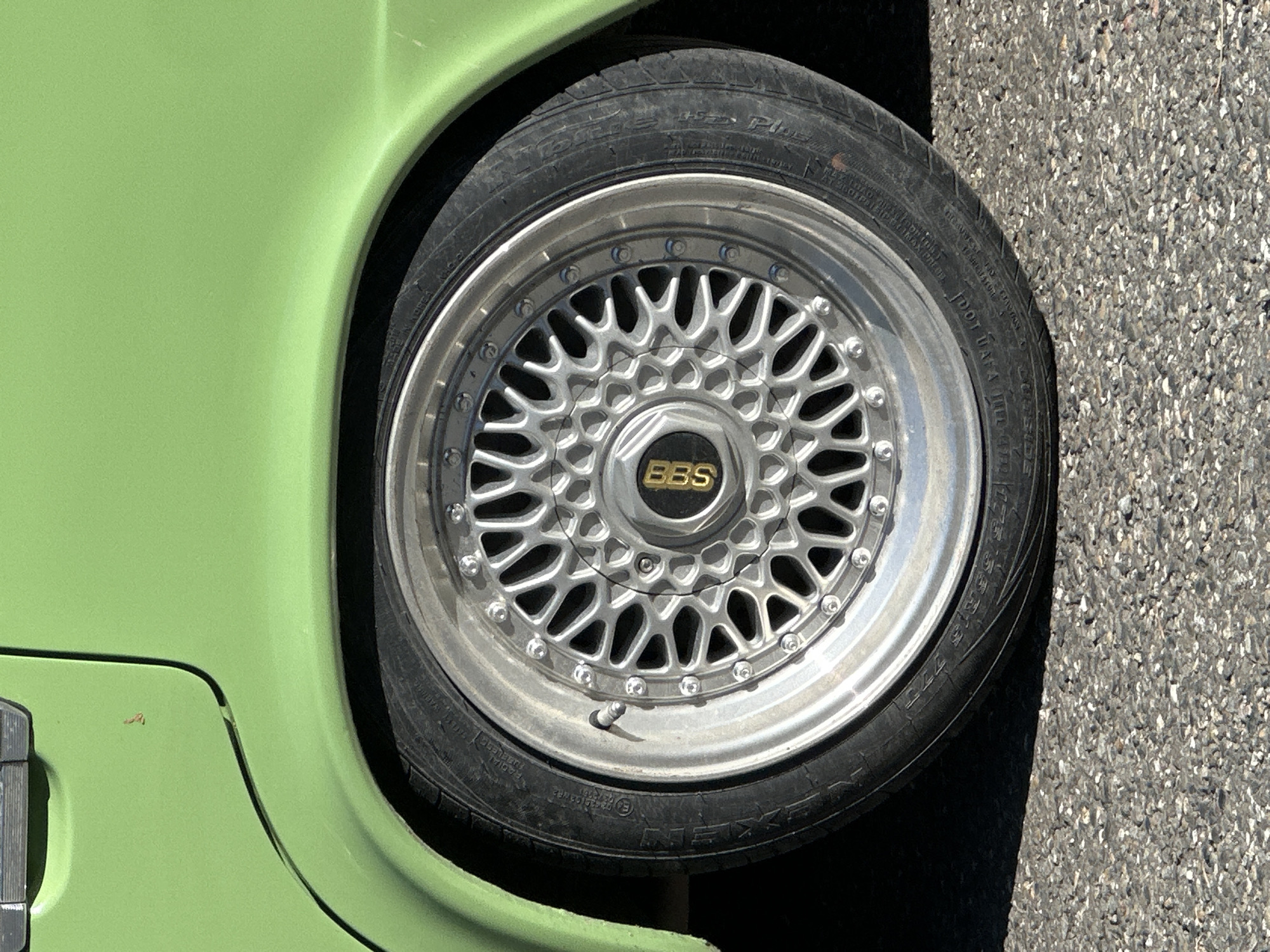 This media file presents a close-up, high-angle shot of the wheel and fender of a vibrant lime green vehicle, parked on an asphalt surface. The car's fender, painted in a bright, almost neon green, curves along the left and top-left edges of the frame. A few minor scuffs or imperfections are visible on the paint near the bottom-left edge of the fender.
The wheel is a silver, multi-spoke design, characterized by an intricate mesh pattern. The center cap prominently features the "BBS" logo in gold lettering on a black background. The wheel's outer rim is secured by numerous small, polished bolts, and a metal valve stem is visible towards the bottom of the wheel. The tire is black and shows visible sidewall markings. Text readable on the tire includes the brand "NEXEN," likely a "PLUS" model, "RADIAL TUBELESS," "DOT UAYA," "OUTSIDE," and the size specification "175/55R15 77S." The tire and wheel appear to have some accumulated dirt and dust.
The ground to the right of the tire is dark, coarse asphalt, indicating a road or parking area. The scene is illuminated by bright daylight, suggesting a sunny day, with subtle shadows providing depth. The image captures a static detail of the vehicle. This image was taken in Krasnaya Polyana, Russia.