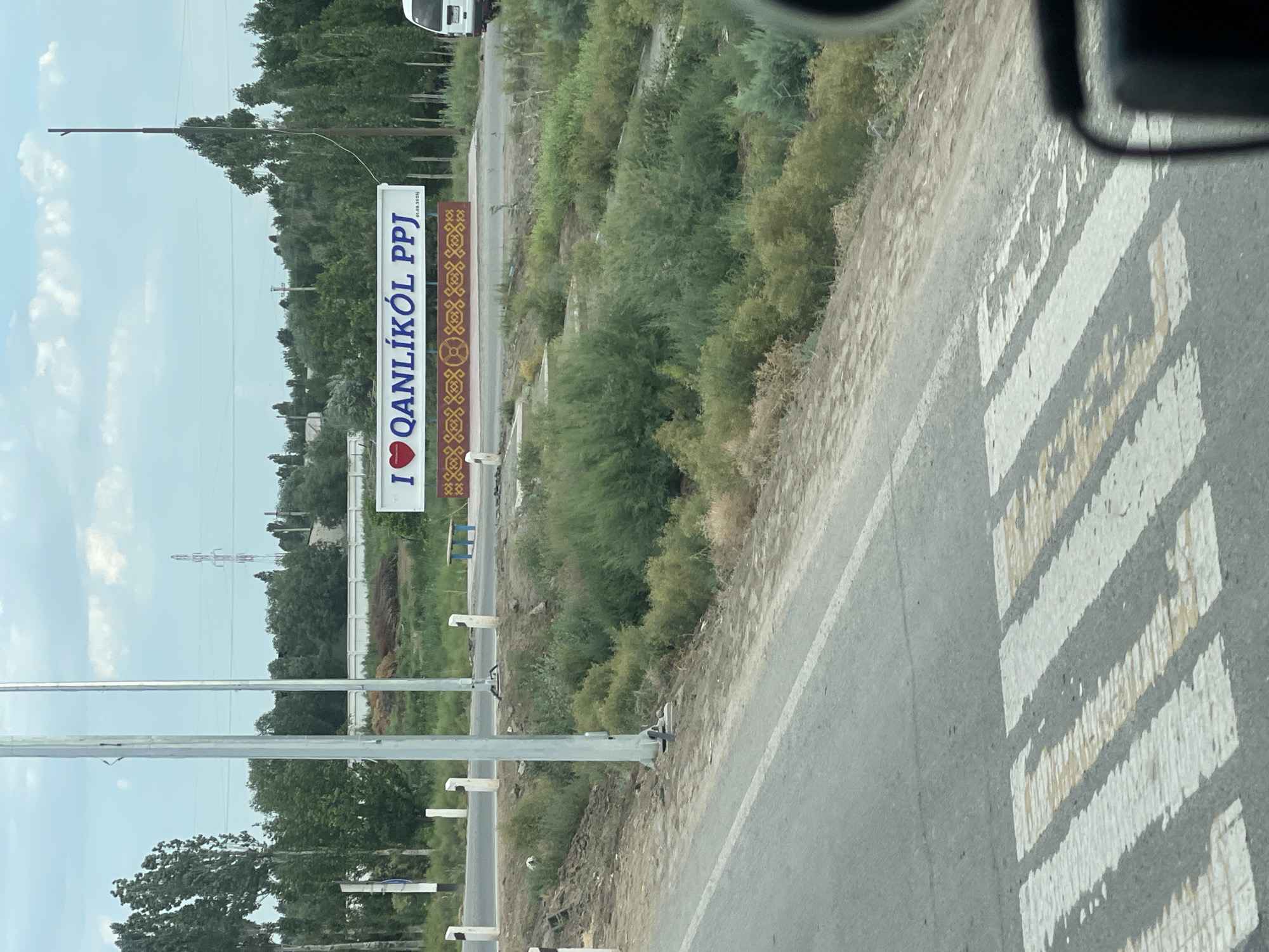 This media file depicts a scene in Qanlikul, Uzbekistan. The primary subject is a roadside sign with bold white text that reads "I ❤️ QANLİKÖL PPJ". The sign also features decorative elements on its right side. The scene appears to be outdoors, likely during the day, with a slightly overcast sky visible. Trees and dense vegetation line the roadside, suggesting a rural or semi-urban setting. A paved road with a pedestrian crossing marked by white stripes is also visible, indicating a public area. The perspective suggests the image was taken from within a vehicle. There are no people clearly visible in the frame. The overall impression is one of a welcoming sign for the region of Qanlikul.