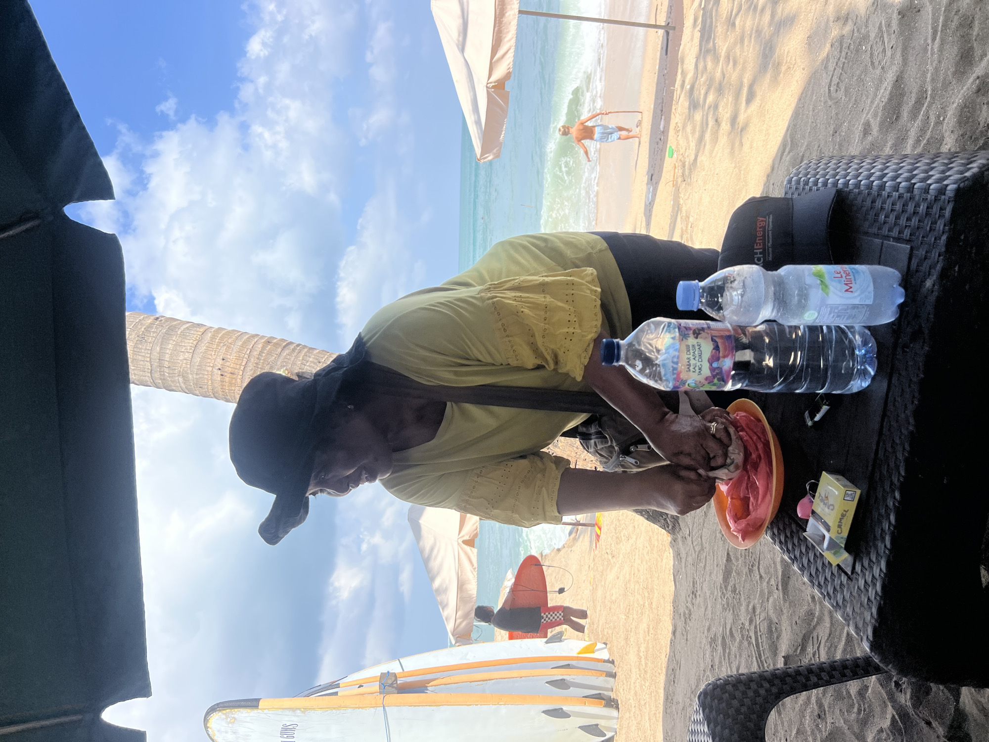 A woman wearing a wide-brimmed hat and a yellow shirt is preparing food on a table at a beach in Kuta, Indonesia. She is seen slicing or preparing something on an orange plate with a pink plastic liner. In front of her on the table are two bottles of water, a pack of Camel cigarettes, and a black baseball cap. Behind her, the ocean waves are visible, with a few people in the water and on the shore. Surfboards are stacked to the left of the frame, and a palm tree is to the right. The scene suggests a casual, tropical beach environment.