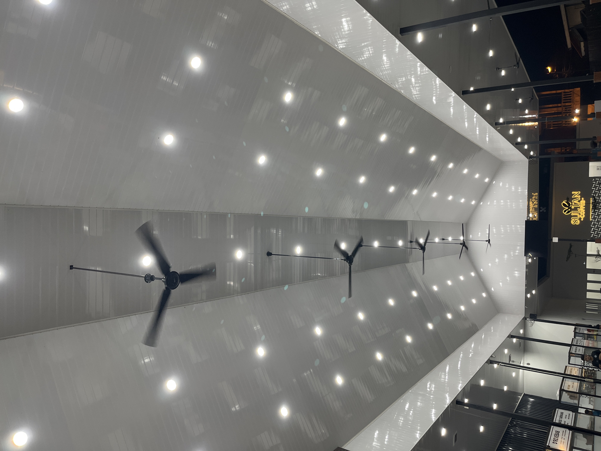 This image captures a wide, modern indoor space, likely a commercial establishment such as a food court or market hall, seen from a low angle looking up towards the high ceiling. The dominant features are the expansive white paneled ceiling, illuminated by numerous bright, circular recessed light fixtures evenly distributed across its surface. Several large, industrial-style ceiling fans are suspended from the ceiling; their blades are blurred, indicating they are in motion for air circulation.
Towards the right side of the frame, a distinct commercial area is visible, featuring various shopfronts or stalls. A prominent lit sign displays the text "SULTAN COFFEE" with a unique logo that resembles two stylized coffee beans or perhaps a bee. Below this, other shop signs are visible, though some text is partially obscured or blurry. One identifiable sign reads "D'RIZ KEBAB" and another seems to say "PINDANG", suggesting a variety of food options.
The overall lighting suggests that the space is well-lit for customers, potentially during the evening hours or in an area without natural light. The architecture is clean and functional, with visible metallic support beams. No people are discernible in the frame, focusing the attention on the structure and amenities of the venue. The scene conveys a sense of a busy, functional, and well-maintained public space in Lhokseumawe, Indonesia.