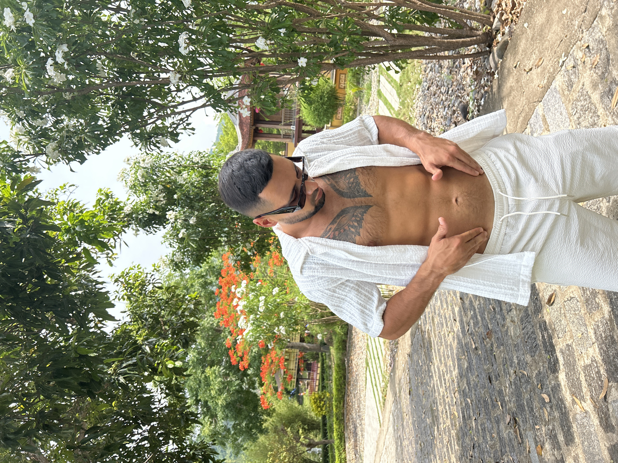 A man stands outdoors, his torso bare and tattooed, wearing a white open shirt and white shorts. He is in a park-like setting with lush green trees and flowering plants. In the background, a traditional Vietnamese-style building with a pagoda roof is visible. The ground is paved with stone and scattered with fallen leaves. The scene appears to be in Nam Nha Trang Ward, Vietnam, given the architecture and vegetation.