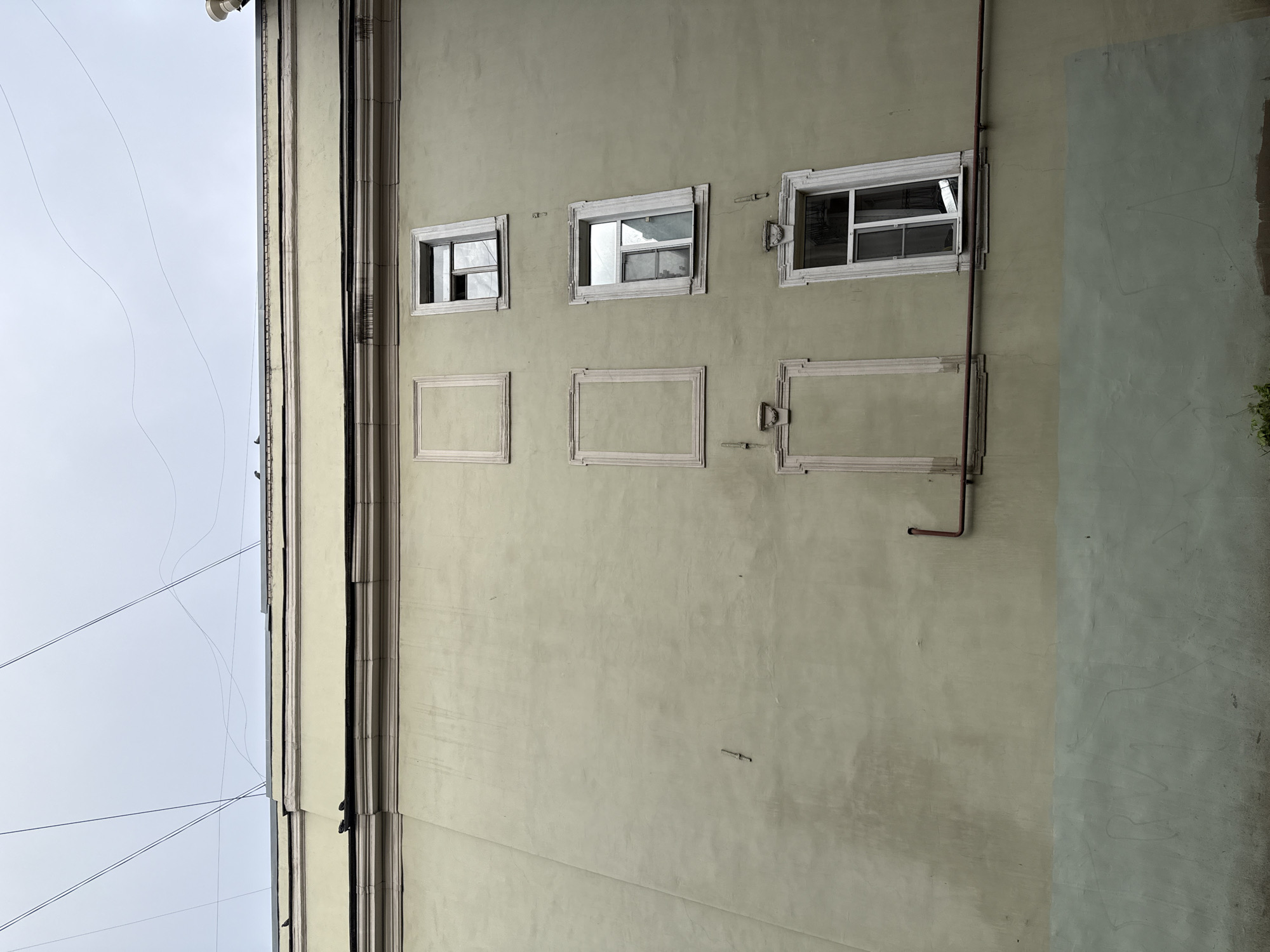 A low-angle, medium shot captures the pale yellow facade of a building in Saint Petersburg, Russia, under an overcast sky. The building features decorative cornices along its upper edge, from which thin black wires are strung across the sky. The wall is punctuated by several windows, some with glass and others appearing as empty frames. Three rectangular frames are present on the left side of the building, one above the other. To the right, three windows are visible in a vertical arrangement. The top window is partially open, revealing a glimpse of its interior. The middle window is also open, with its panes angled. The bottom window is closed and appears to be a standard window frame. A rusty pipe runs horizontally across the lower section of the facade. The lower part of the wall transitions to a muted green color, with faint scrawled markings on its surface. Small birds are perched on the architectural elements of the building.