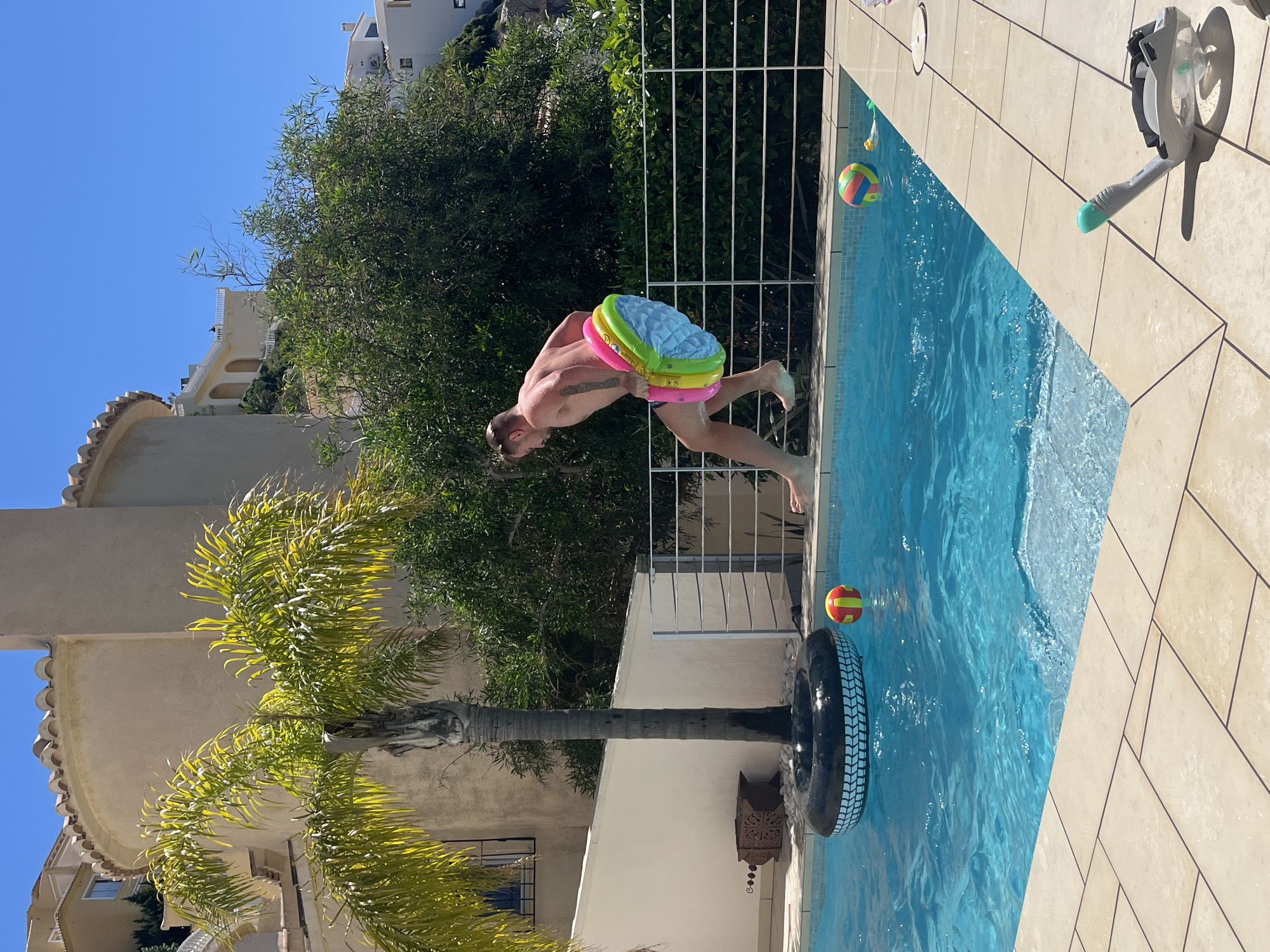 A medium, eye-level shot reveals a person mid-leap into a swimming pool at a location identified as el Poble Nou de Benitatxell / Benitachell, Spain. The person, a muscular male, is airborne with a multi-colored inflatable seat pad positioned around his waist.
The pool, filled with clear blue water, features a large black tire floating in it, with two multi-colored balls present. The poolside is paved with beige-colored tiles. A full-face snorkel mask rests on the pool deck, partially out of frame. A white metal fence separates the pool area from a lush green shrubbery, behind which the exterior of a building can be seen. A palm tree stands at the edge of the pool area. The sky is clear and blue.