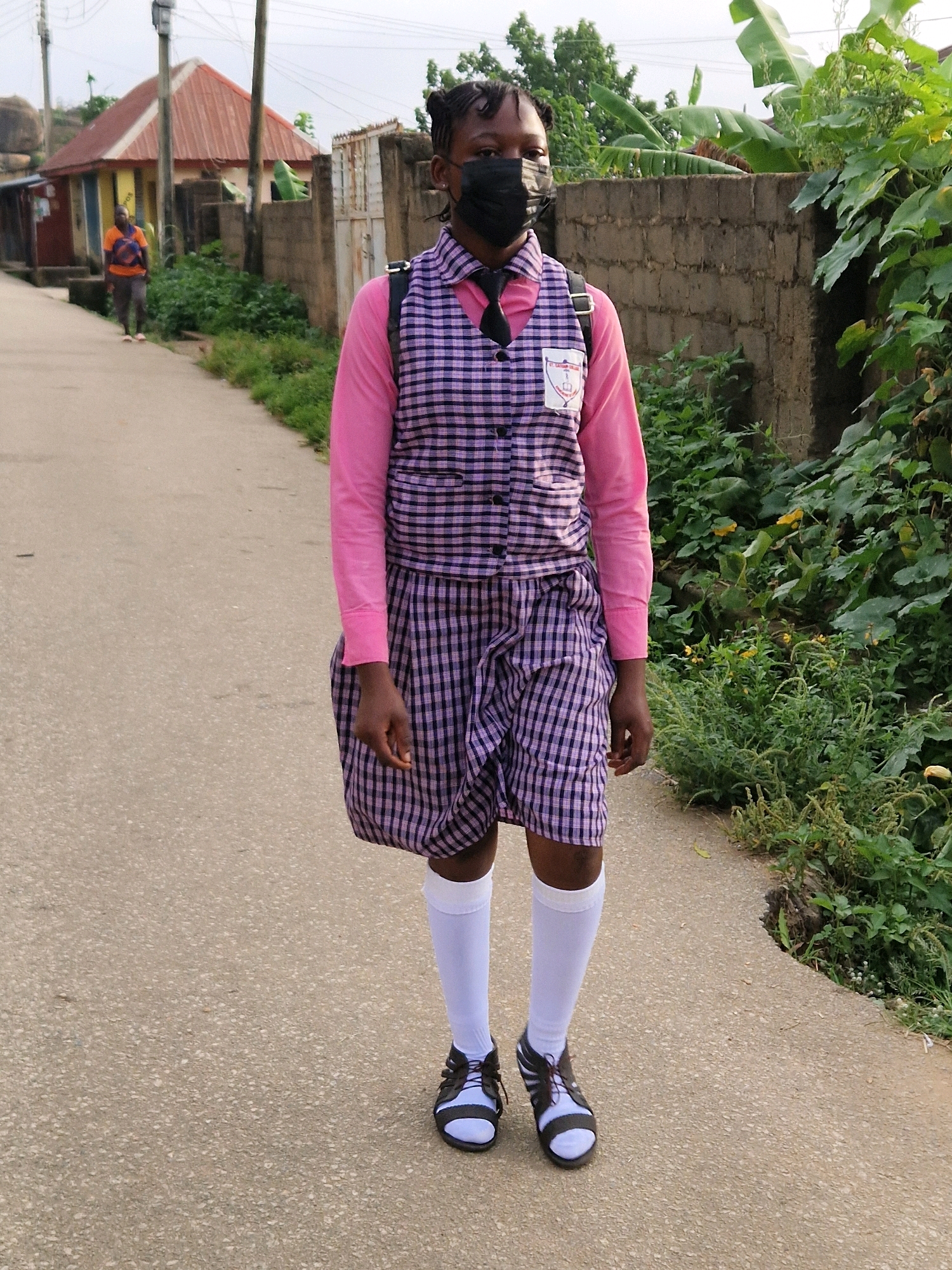 A young Black girl wearing a school uniform, consisting of a pink long-sleeved shirt, a checkered vest and skirt, white knee-high socks, and black and white sandals, walks on a paved path in an outdoor setting. She has dark hair braided and pulled back, and is wearing a black face mask. A black backpack is visible on her shoulders. Behind her, the path continues, bordered by a concrete wall covered in vegetation on the right and some buildings on the left. The buildings appear to be a mix of residential structures with various roof colors, including red and orange. In the background, a person wearing an orange shirt and gray pants is walking away from the camera. The overall environment suggests a residential area in Jos, Nigeria, with a slightly overcast sky.