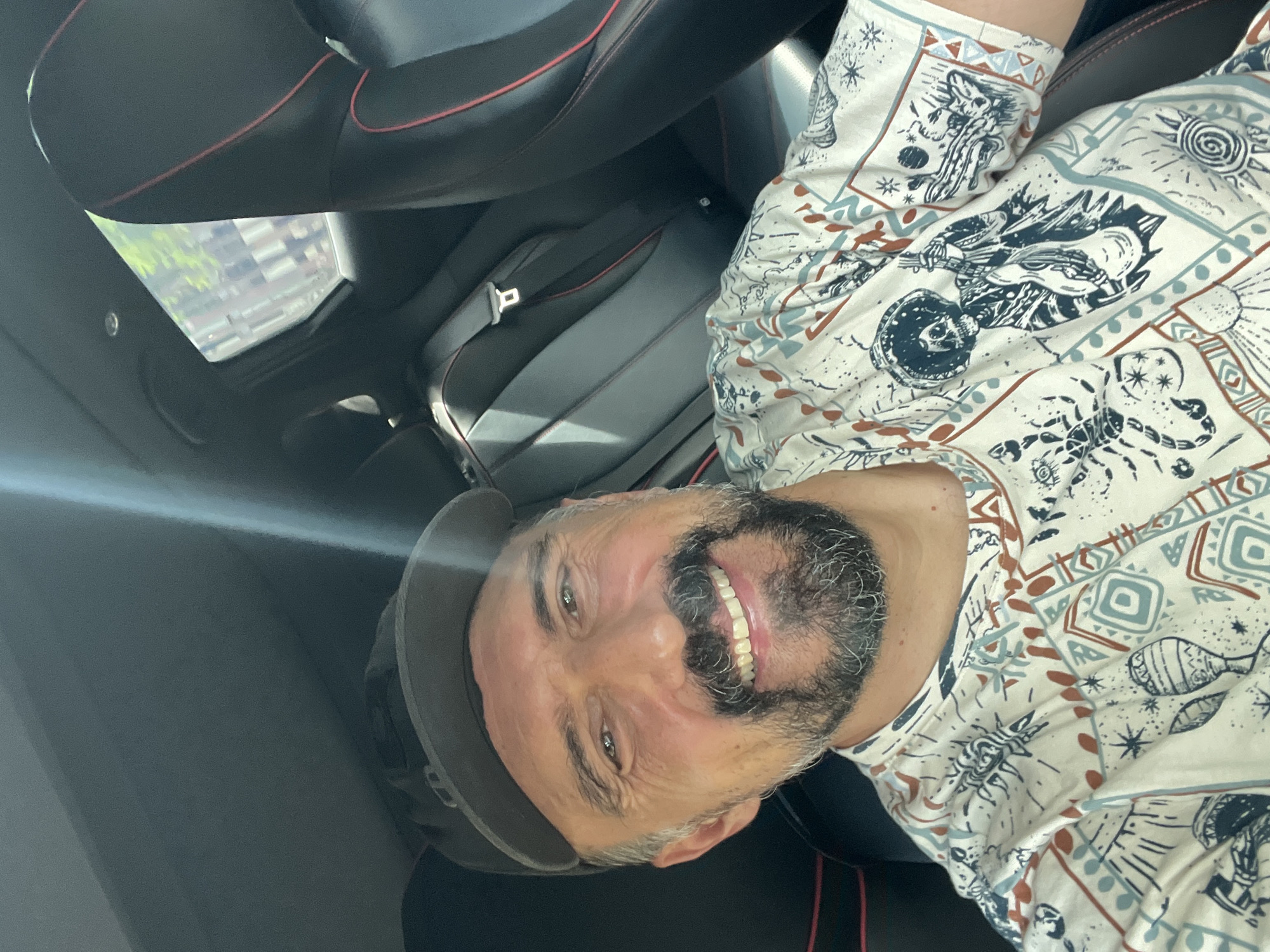 A man wearing a black baseball cap and a white t-shirt with a skull and guitar pattern is taking a selfie in the driver's seat of a car. The car's interior is visible, with black leather seats accented with red stitching. Sunlight streams in from the side, creating a glare on the windshield. Through the window, a blurry view of buildings and trees can be seen, suggesting an urban or suburban environment. The man has a beard and is smiling. The image is taken outdoors, likely during the day, given the bright lighting.