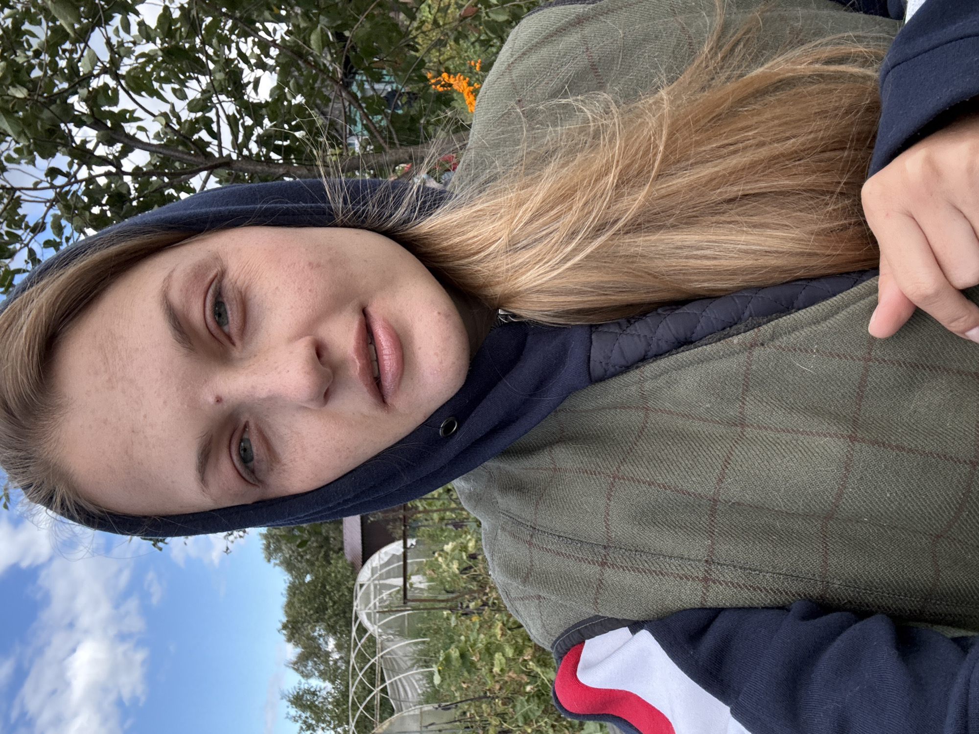 A woman with blonde hair, wearing a dark blue hoodie and a green plaid vest over a black quilted jacket, stands outdoors in Fominka, Russia. Her left arm is visible, with her hand resting near her chest. The background shows greenery, including leafy trees and some type of agricultural structures resembling greenhouses. The sky is partly cloudy.