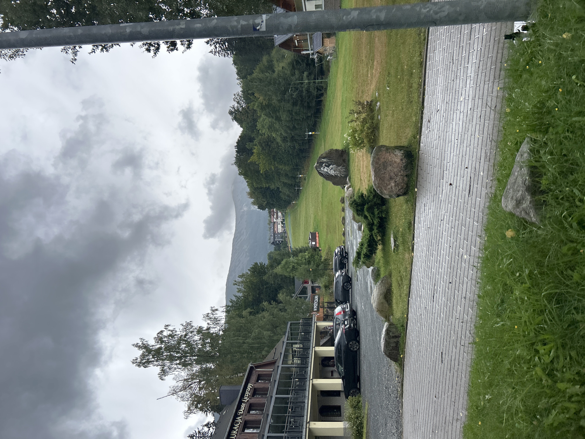 The image depicts a cloudy, overcast day in Karpacz, Poland, likely at a ski resort or a mountain lodge area. In the foreground, a paved walkway with a brick pattern leads to a grassy area with several large rocks and low-lying bushes. To the left, a multi-story building with a dark wooden exterior and large glass windows, identified by signage as "RESTAURACJA DWÓR LICZYRZEPY", is visible. Several cars are parked in a gravel lot in front of this building, including a black sedan, a dark grey SUV, and two other dark-colored vehicles.
In the midground, a grassy slope rises, dotted with scattered rocks and small trees. A large boulder prominently features a painted image of a bearded face. A ski slope or hiking trail is suggested by the presence of yellow poles with signage. Further back, a dense forest of green trees covers the hillside, with some buildings and structures peeking through the foliage. The background shows more of the forested mountain landscape, with the peaks partially obscured by clouds. The overall atmosphere is damp and cool, consistent with a mountainous climate on a cloudy day. A vertical metal pole, possibly a lamppost or signpost, is present on the right side of the frame, partially obscuring a section of the scene.
