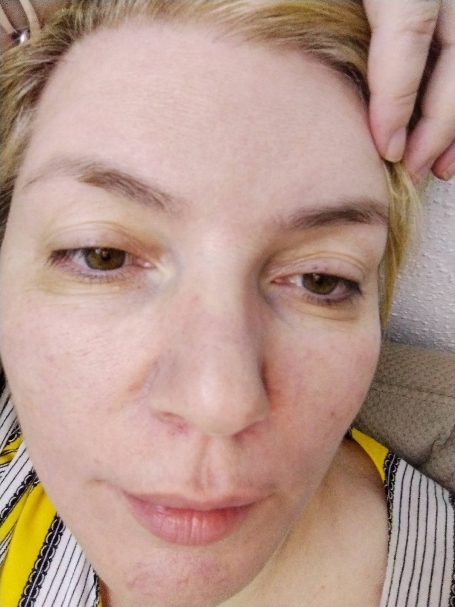 A close-up, selfie-style portrait of a fair-skinned woman with blonde hair, visible from the forehead to the upper chest. She has brown eyes and is looking slightly downward. A hand is partially visible at the upper right of the frame, pulling at the hairline. The woman is wearing a garment with a yellow and white striped pattern. The location of Al Mas'oudi, United Arab Emirates is not evident from the image.