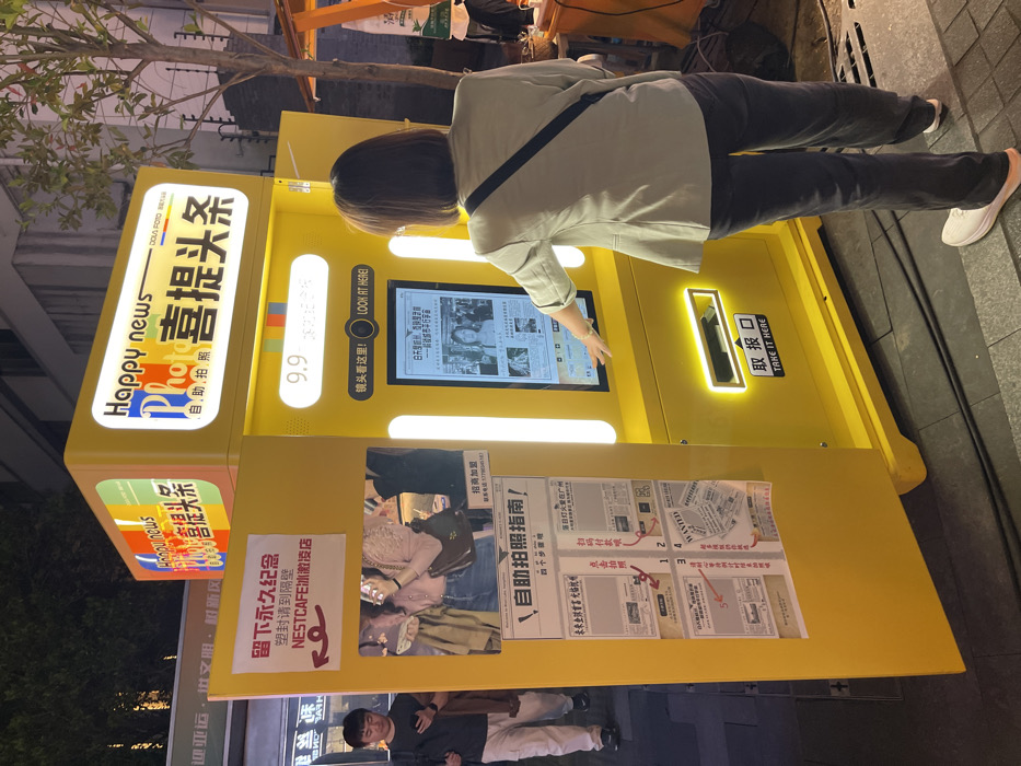 The image shows a yellow photo booth, located on a street in Hangzhou Shi, China at the coordinates 30.263 latitude and 120.156 longitude. The photo booth is labeled "Happy News Photo Booth" and is adorned with Chinese and English text. A woman is interacting with the machine, possibly paying for or selecting a photo option. The screen displays a headline for a news article, visible behind the woman. The photo booth is situated on the sidewalk next to a building. The pavement is clean and a black cable snakes across the ground. The photo booth has a large yellow door open, revealing a sign with Chinese characters. To the left of the photo booth, a man stands with a backpack on, looking down at his phone. Behind the booth, the street is dimly lit, suggesting it is nighttime. A sign hanging above the booth's door reads "NestCafe." To the left of the photo booth, a sign that says "Welcome to Nemcafe" is visible. The scene suggests a bustling city environment.