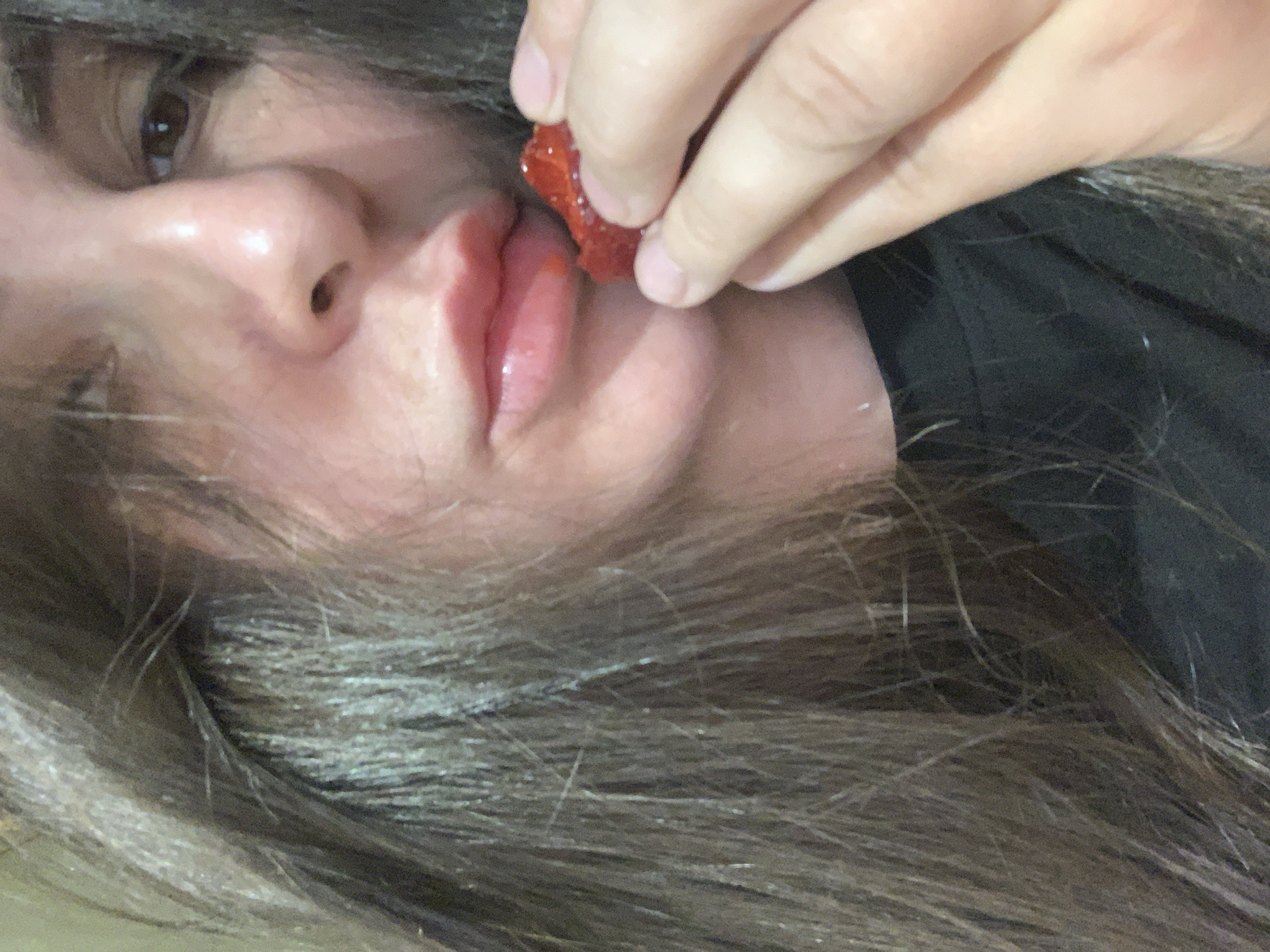 The close-up shot captures a person's face, with their hair framing the left side of the image. The subject is in the process of eating a strawberry, with their fingers holding the fruit to their lips. The lighting is soft, highlighting the texture of the skin and the sheen on the lips. The background is indistinct, suggesting a shallow depth of field. The overall impression is one of a candid moment, possibly taken in a private setting.
