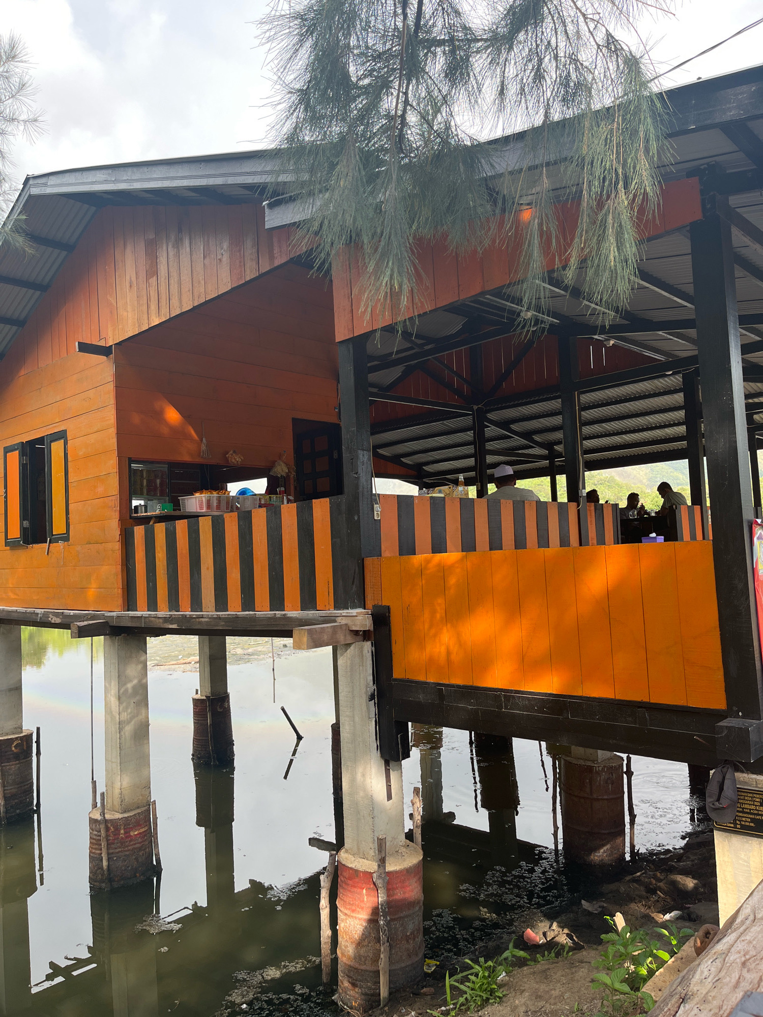 The image depicts a stilt house on a body of water in Aneuk Paya, Indonesia. The house features orange and black striped railings and wooden walls. Several people are seated inside the open-air structure, appearing to be dining. The stilts supporting the house are made of concrete and rusted metal barrels. The water is murky and appears to have some debris. The surrounding environment is natural with trees and a cloudy sky. A small rainbow can be seen in the water reflecting off the stilts.