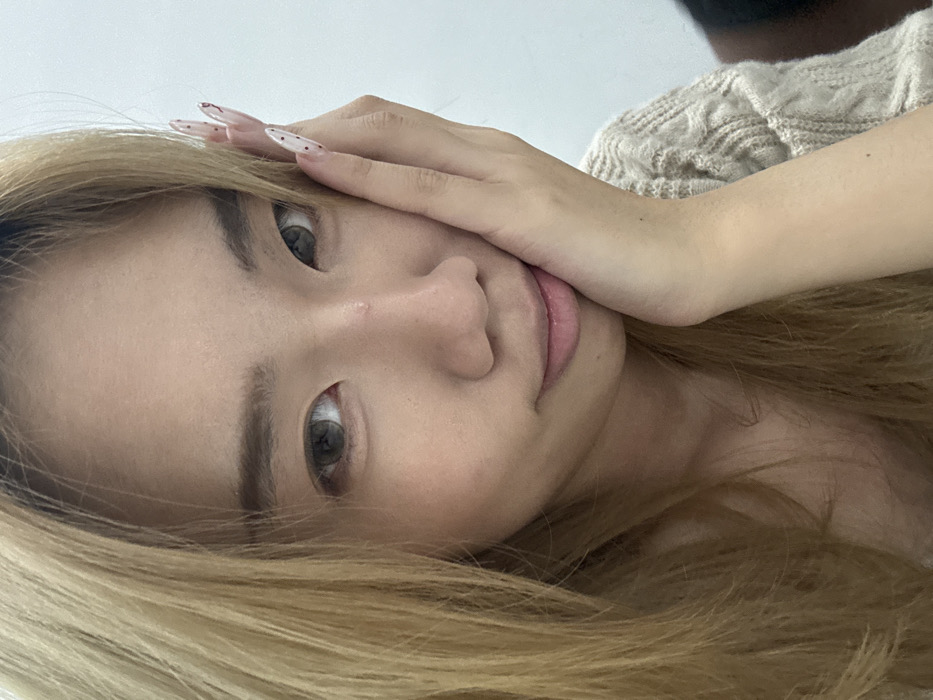 A close-up portrait features an East Asian female visible from the chest up. She possesses long, light blonde hair, parted to her right, with bangs framing her face. Her eyes, appearing to have greyish-brown contact lenses, are directed towards the viewer. A small blemish is visible on the bridge of her nose. Her lips are slightly parted, suggesting a subtle smile. Her left hand rests against her left cheek, with the palm touching the skin and fingers extending upwards. Her fingernails are long, pointed, and covered in clear polish featuring small dark red or brown polka dots. She wears a light-colored, possibly beige, cable-knit sweater with a visible pattern. The background is uniformly plain and light-colored, providing no discernible architectural or environmental details to establish a specific location within Yuzhong District, China.